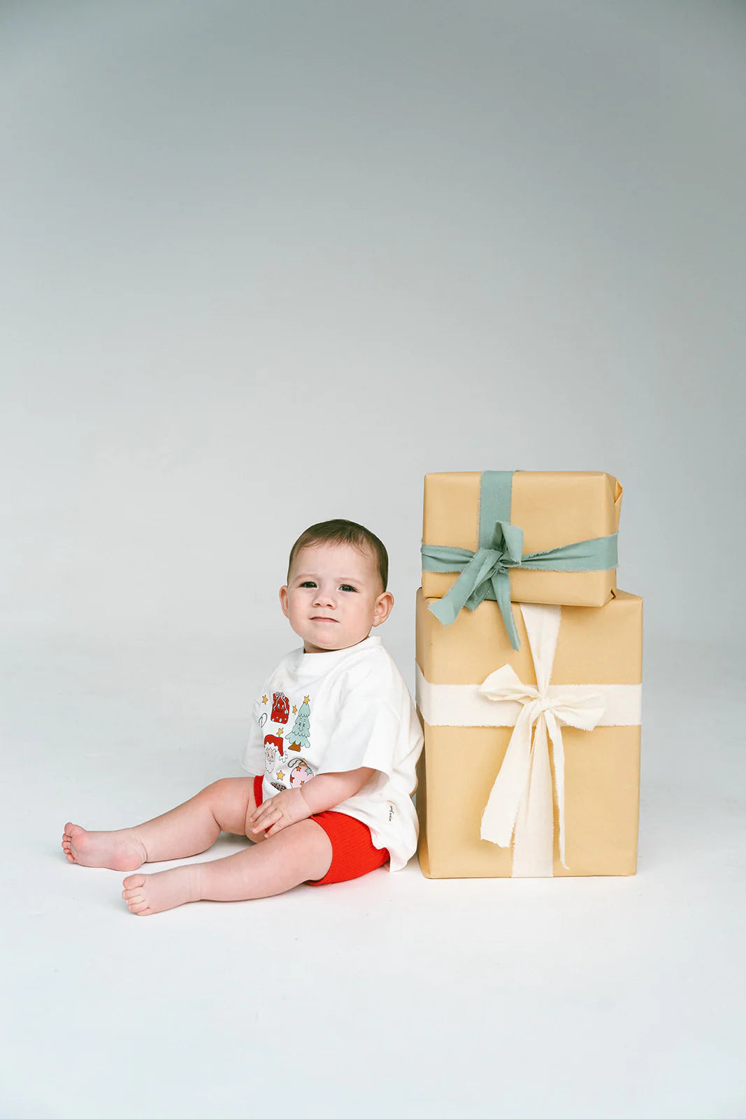 Wearing a Merry & Bright Icons Oversized Tee by MYLA JANE, the baby sits on the floor next to two large wrapped gifts adorned with bows, perfectly embodying the holiday season spirit. Set against a plain light gray backdrop, the baby's neutral expression adds charm to this festive scene.