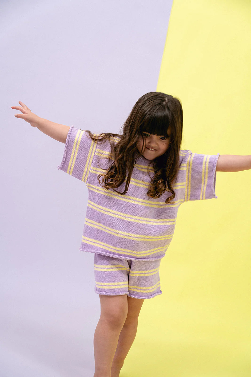 A young girl with long brown hair joyfully balances with outstretched arms, showcasing the Contrast Knit Set Lemon/Grape from MYLA JANE. She is wearing a purple and yellow striped ensemble against a pastel backdrop.