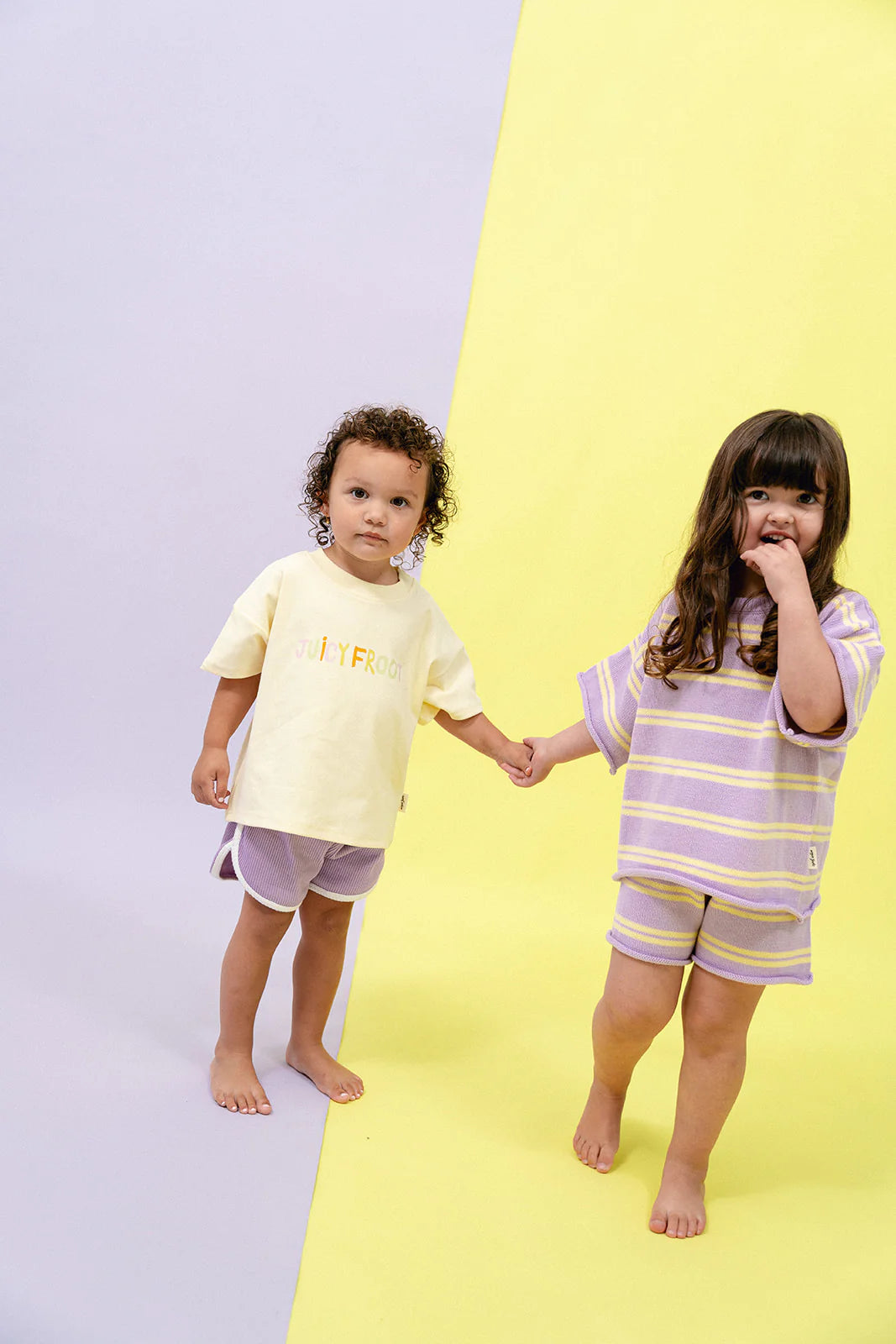 Two children stand hand in hand, perfectly capturing the playful essence of the MYLA JANE Juicy Froot range. The child on the left wears the Contrast Knit Set Lemon/Grape, showcasing a light yellow shirt paired with purple shorts. Simultaneously, the child on the right sports a vibrant combination of purple and yellow stripes. Their vivid attire pops against a backdrop echoing their mix-and-match style.