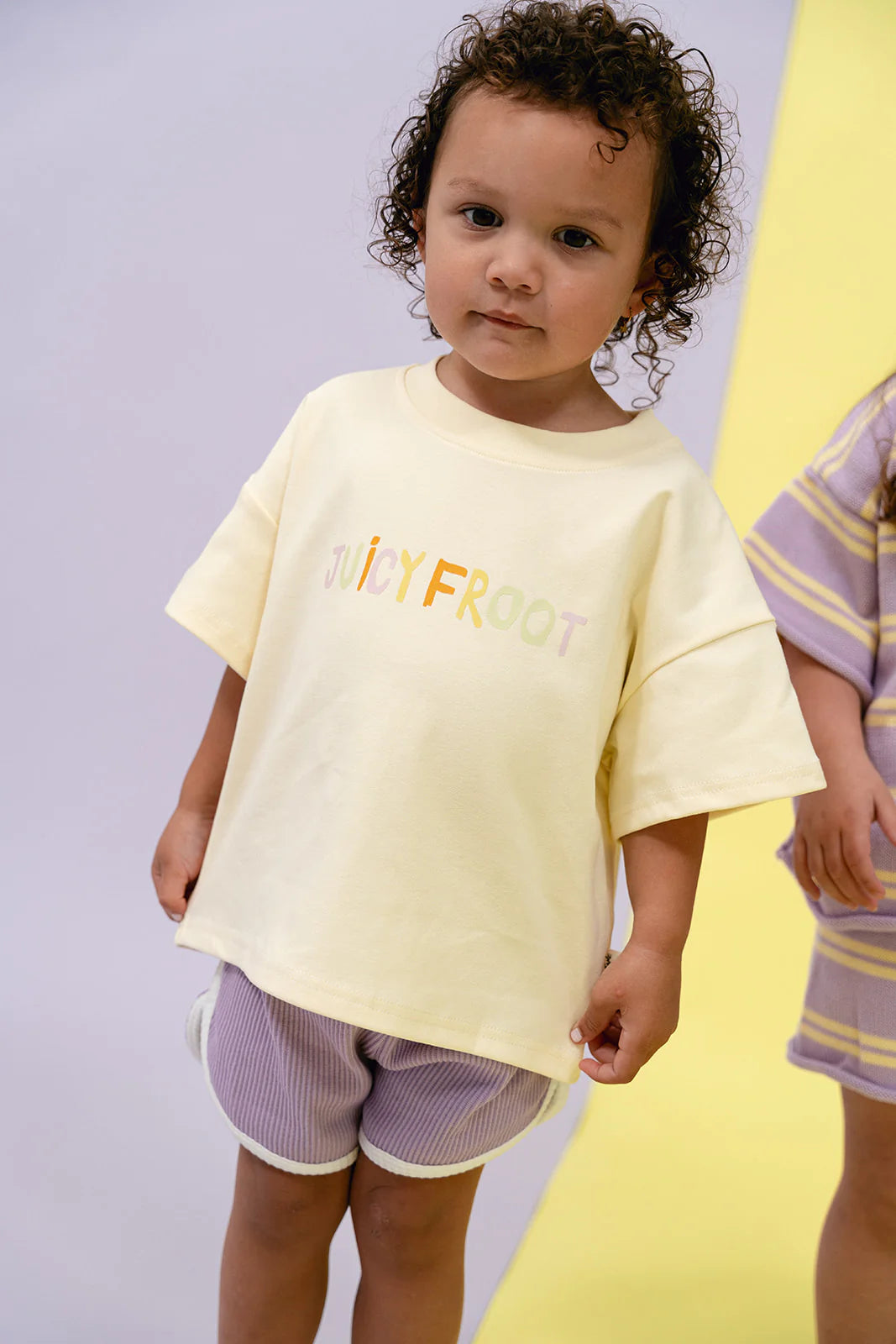 A young child with curly hair sports a light yellow Juicy Froot Tee paired with grape-colored MYLA JANE Contrast Rib Shorts. They pose on a backdrop divided between white and yellow, alongside another child in similar oversized relaxed design shades partially visible next to them.