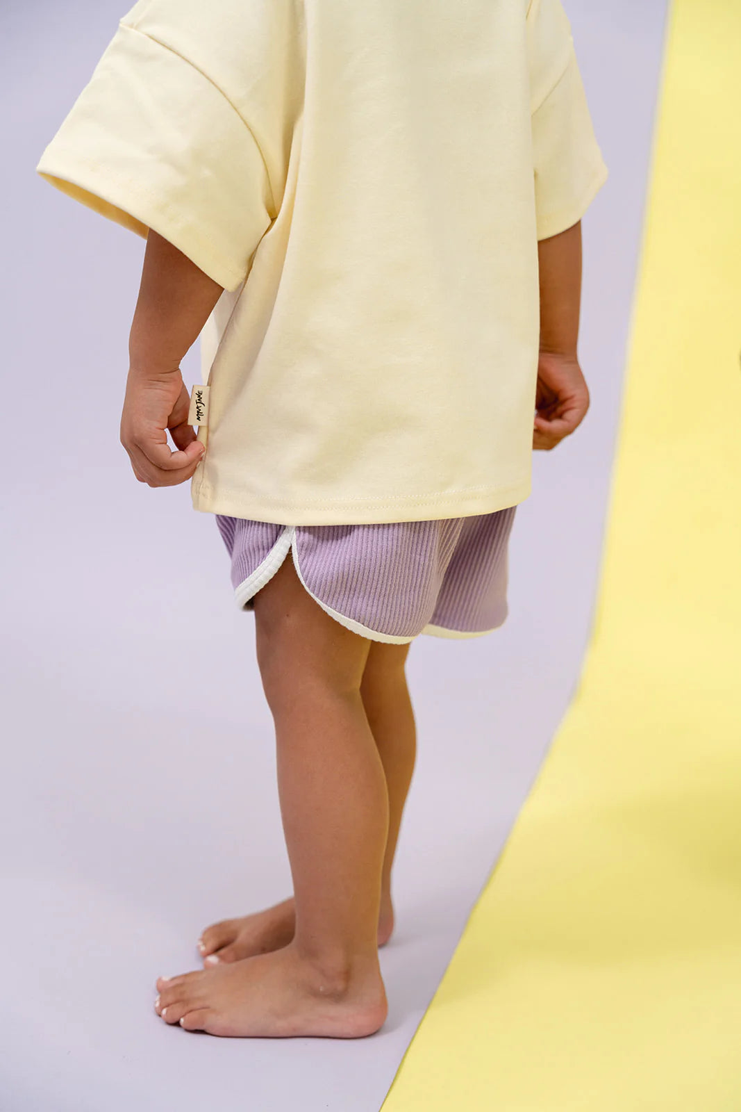 A child in an oversized, relaxed cream-colored T-shirt and vibrant MYLA JANE Contrast Rib Shorts in Grape stands barefoot, with one foot on a gray surface and the other on a yellow surface. The image focuses on the child's legs and torso, capturing the playful essence of this attire.