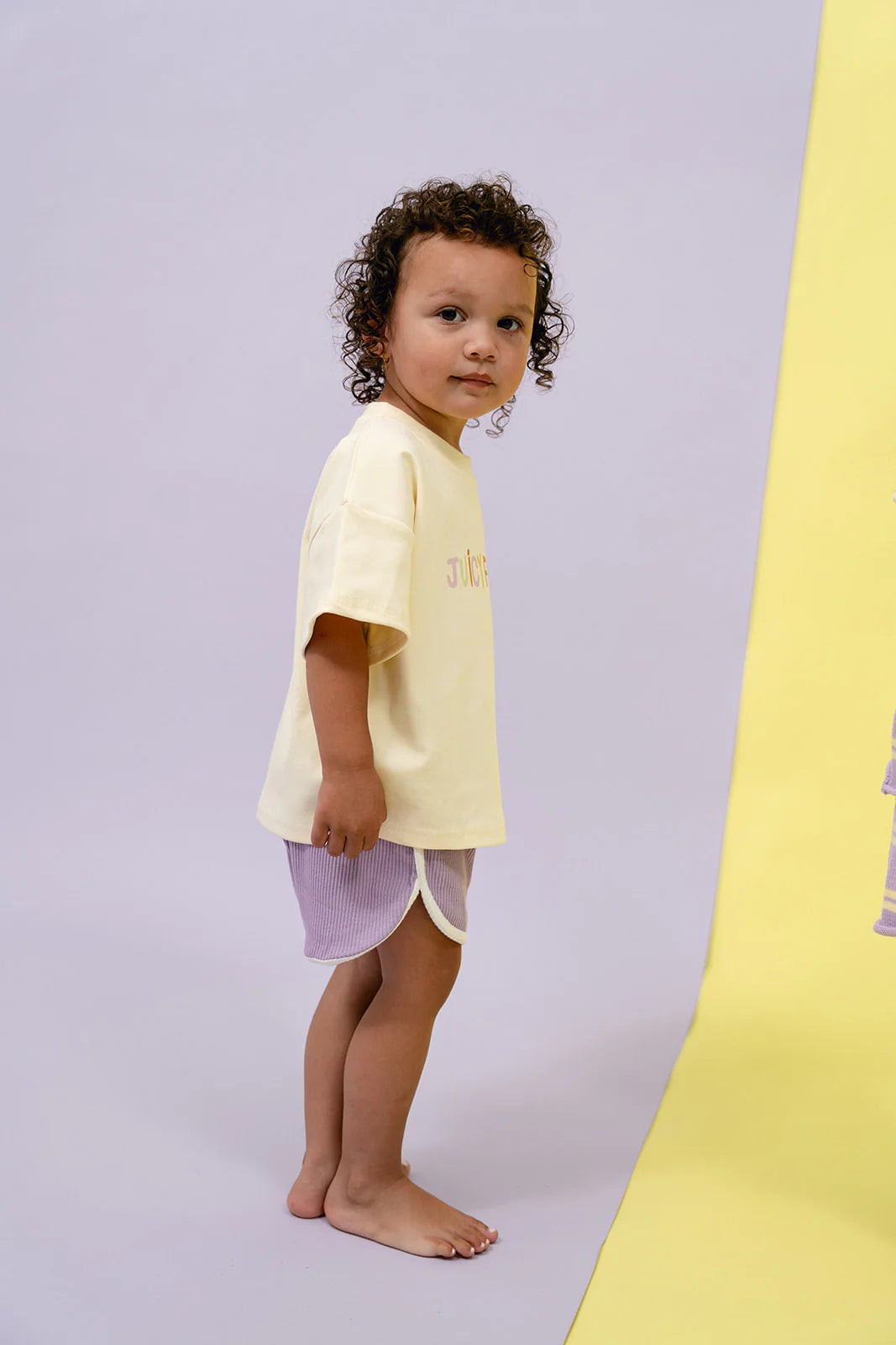 A young child with curly hair stands barefoot against a light-colored backdrop. Sporting an oversized relaxed design, the child is wearing a yellow Juicy Froot Tee paired with vibrant MYLA JANE Contrast Rib Shorts in grape, looking slightly to the right.