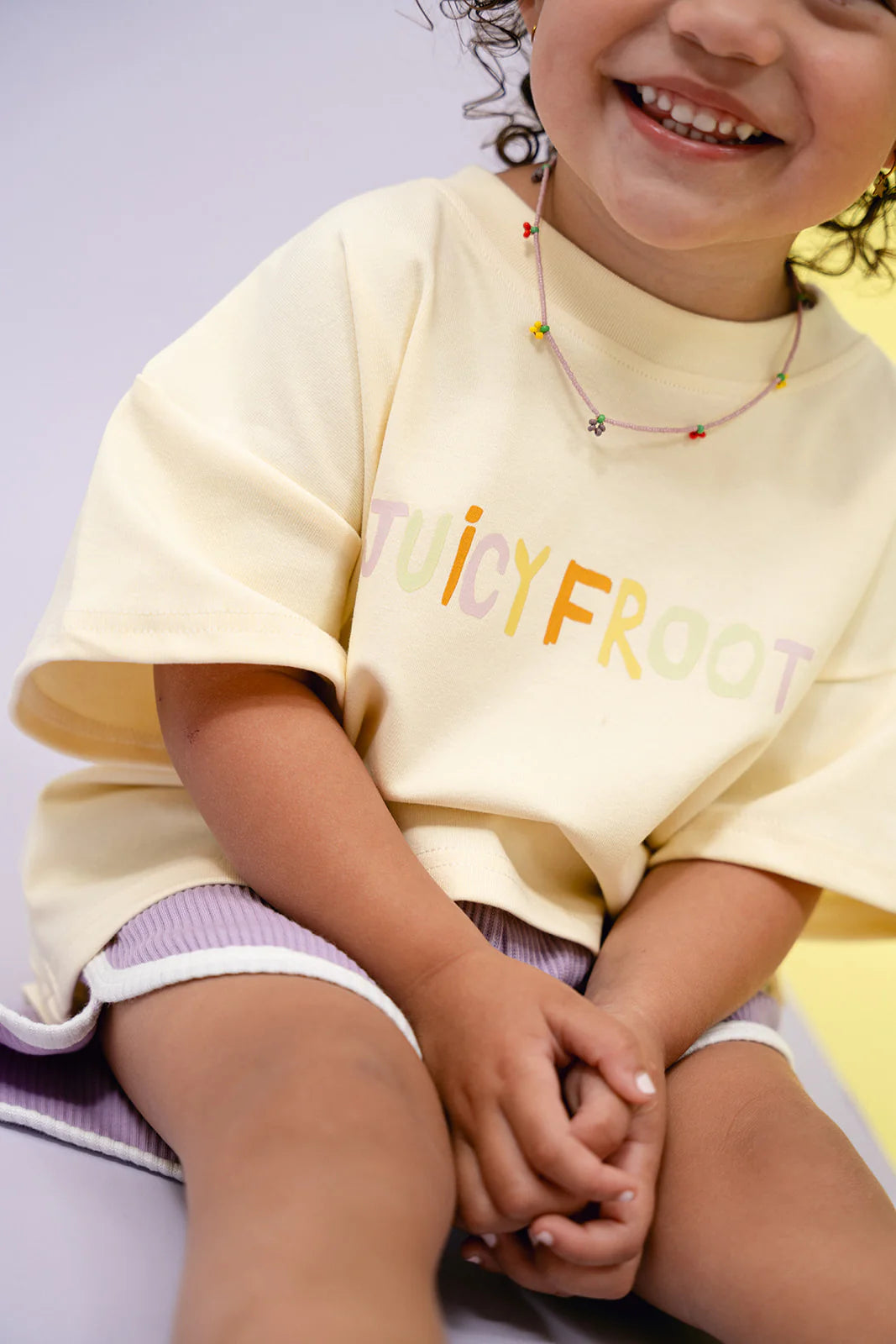 A young child with curly hair smiles while sitting, wearing a Juicy Froot Tee and MYLA JANE's Contrast Rib Shorts Grape. Their yellow t-shirt displays "JUICY FROOT," and their purple shorts, trimmed in white, offer a vibrant pop of color. A small beaded necklace completes the look against the softly lit pastel background.