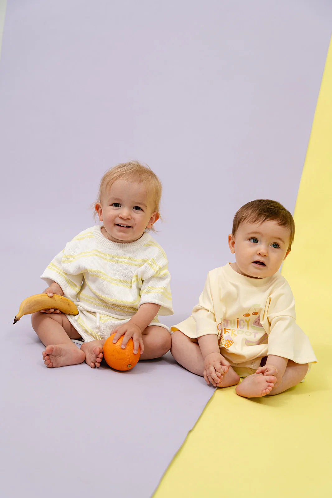 Two toddlers sit on a dual-colored backdrop; one holds a banana while the other clutches an orange. They are dressed in the MYLA JANE Contrast Knit Set Lime/Off White, crafted from 100% cotton for perfect mix-and-match attire. Both are barefoot and gaze curiously at their surroundings.
