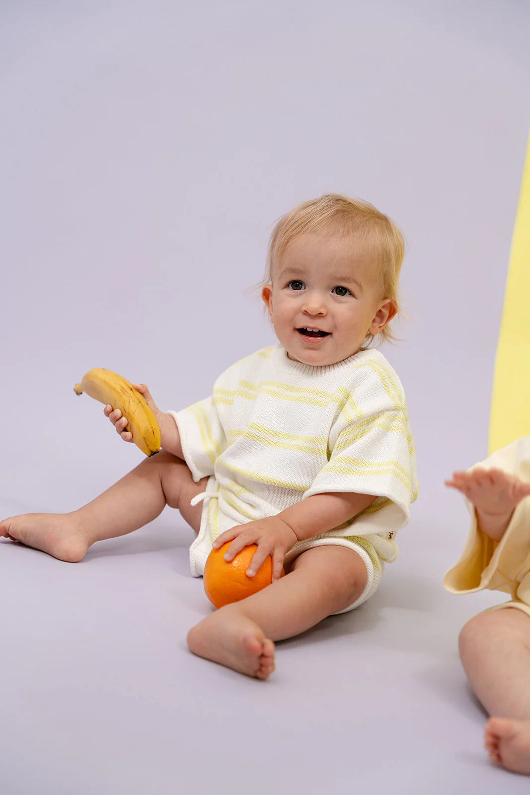 A toddler sits on a gray surface, holding a banana in one hand and an orange in the other, wearing the MYLA JANE Contrast Knit Set Lime/Off White, which is crafted from 100% cotton. The child looks at the camera while another child is partially visible beside them, both enjoying the versatility of mix and match styles.