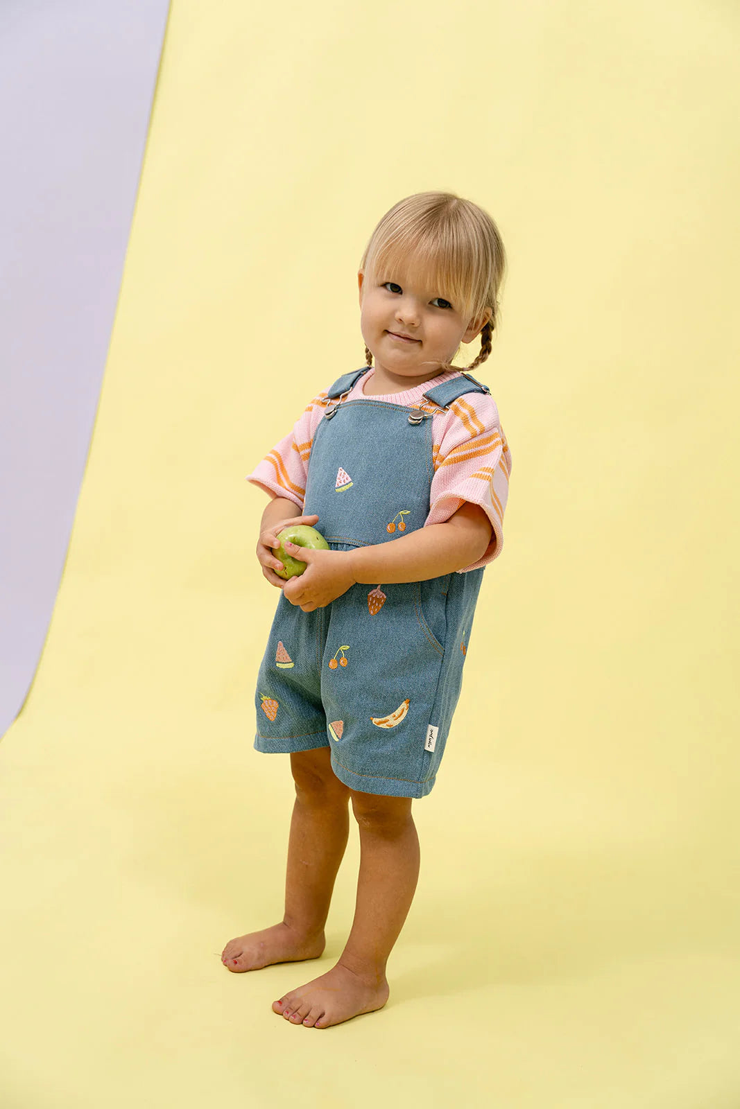 A toddler with blonde hair stands barefoot on a yellow background, wearing a MYLA JANE Contrast Knit Set in Strawberry/Orange, holding a green apple.