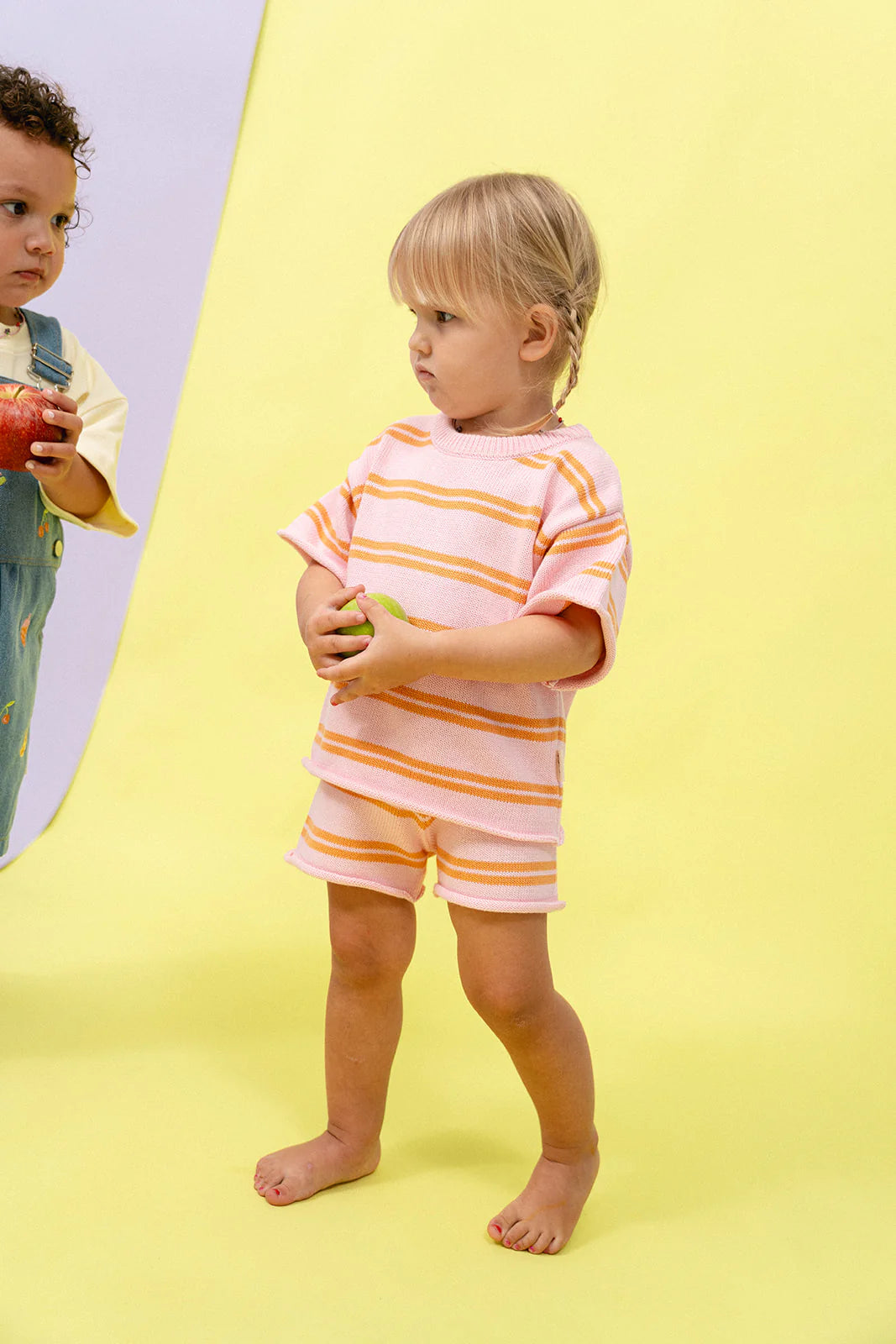 A young, blond-haired child, barefoot on a yellow background, wears the Contrast Knit Set Strawberry/Orange from MYLA JANE and holds a green apple. Nearby, another child in a Juicy Froot-themed blue outfit with colorful prints clutches a red apple.