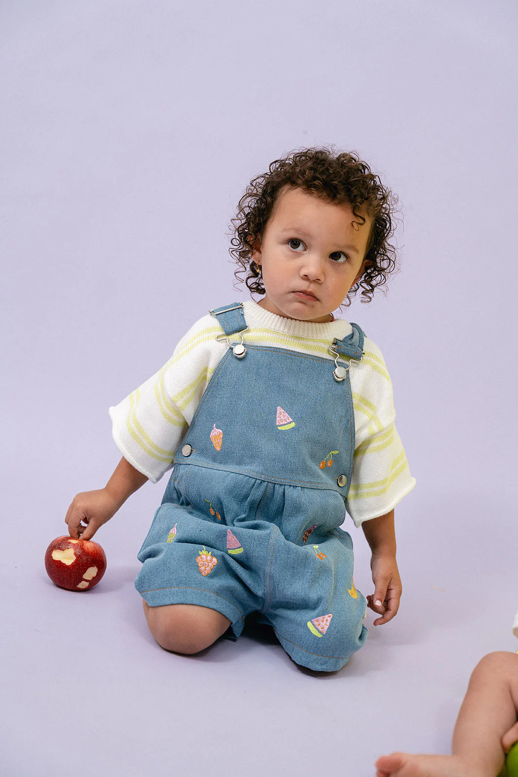 A toddler with curly hair sits on the floor, dressed in the Contrast Knit Set Lime/Off White from MYLA JANE, featuring a striped top and denim overalls adorned with fruit designs, crafted entirely from 100% cotton. They are holding an apple with a heart-shaped piece taken out of it, set against a soft pastel background.