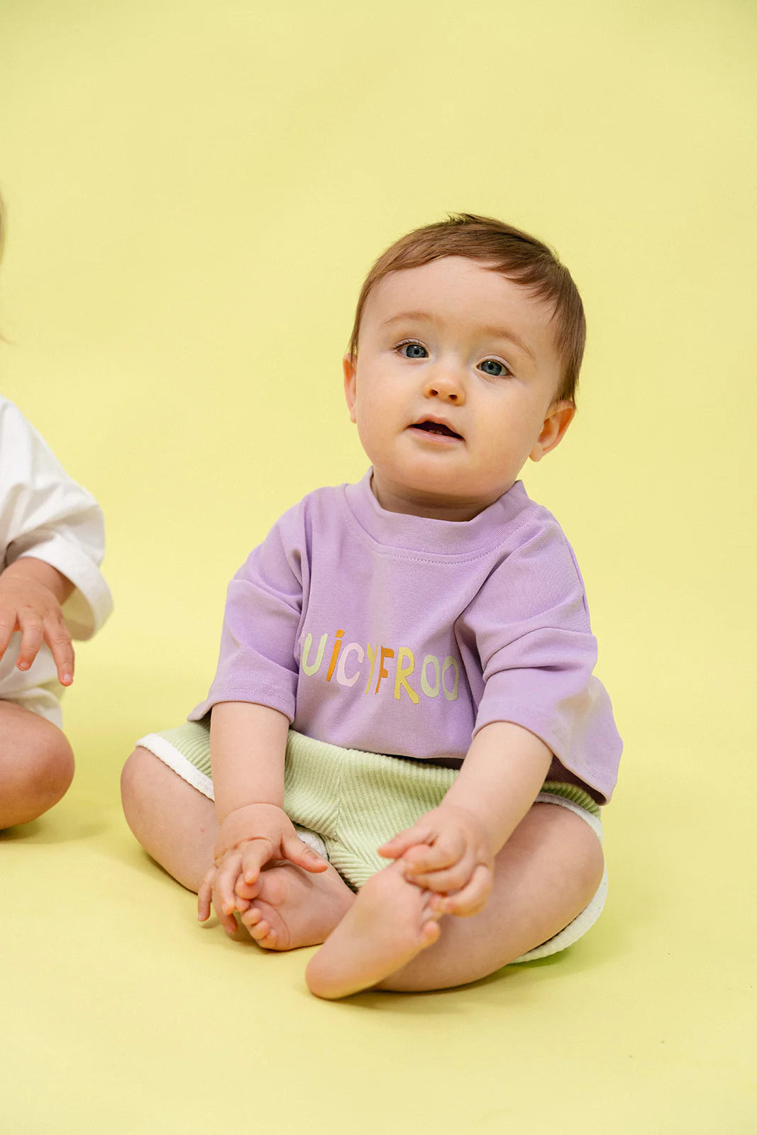 A baby sits on a yellow background, wearing an oversized purple shirt with "JUICYFRUIT" emblazoned on it and vibrant Contrast Rib Shorts in lime by MYLA JANE, their neutral gaze directed upwards.