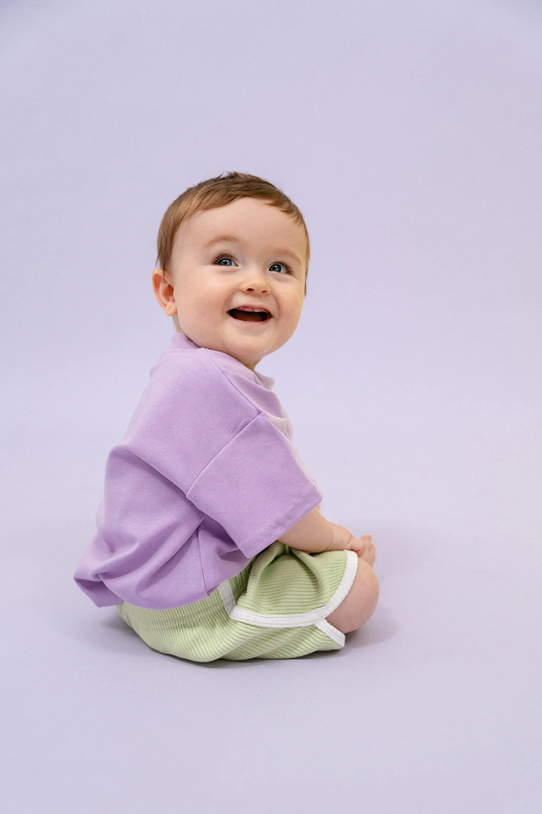 A smiling baby sits on a light purple background, wearing a lavender shirt made from soft cotton rib. The outfit is completed with MYLA JANE's lively Contrast Rib Shorts in Lime, which feature striking white stripes. The baby, with short brown hair, gazes upward and to the side.