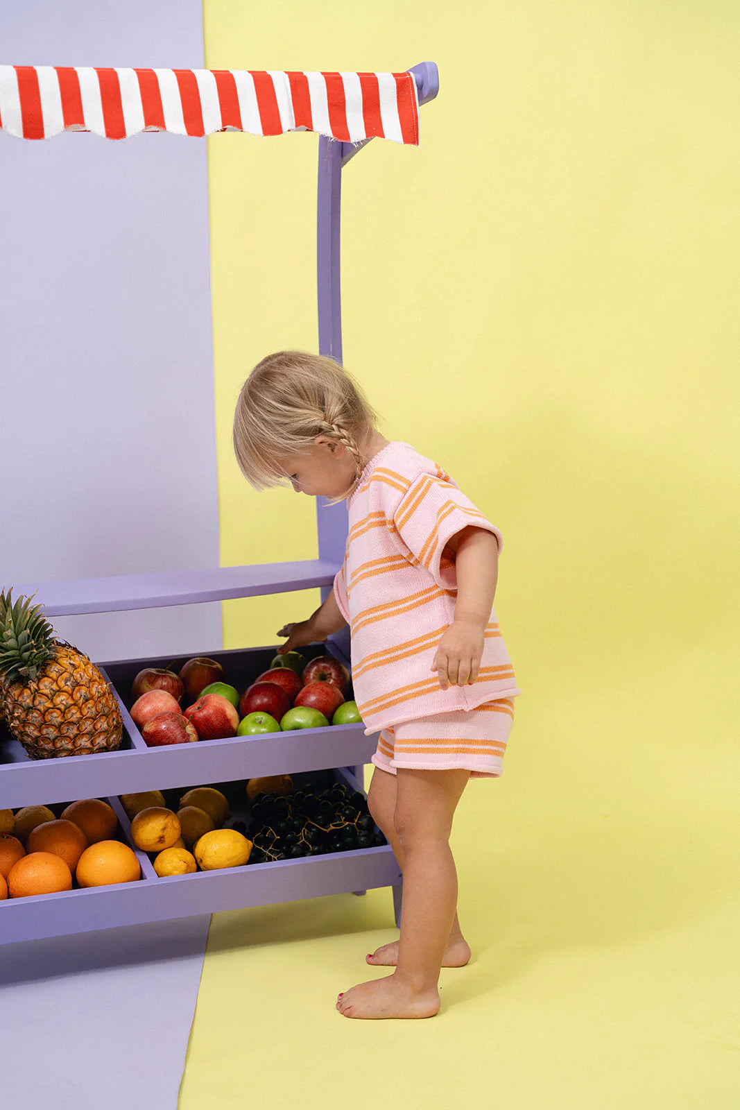 A toddler wearing the Contrast Knit Set in Strawberry/Orange by MYLA JANE stands beside the Juicy Froot cart. The cart, featuring a red and white striped canopy, is brimming with pineapples, apples, oranges, and grapes against a backdrop of yellow and light purple.