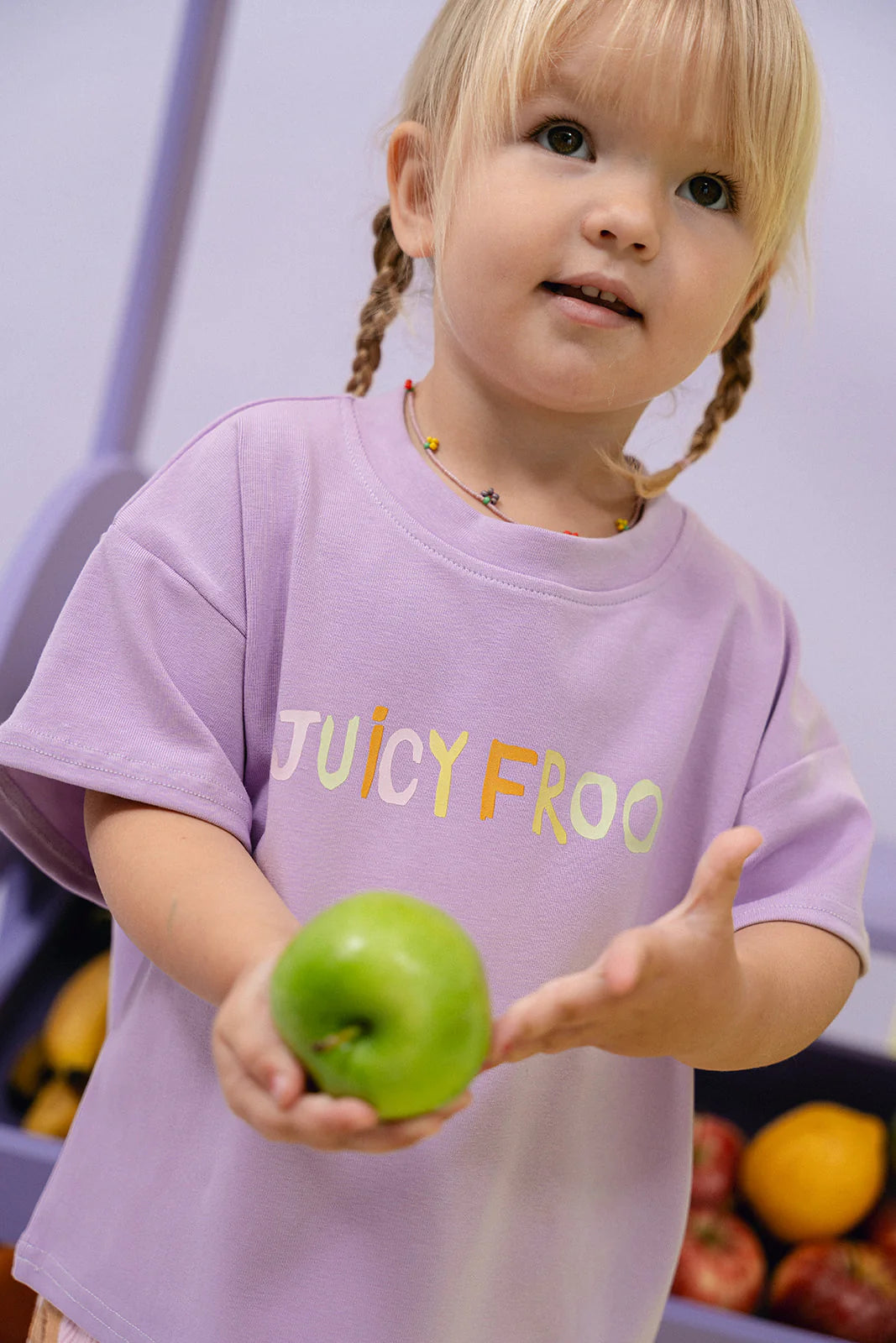 A young child with blonde braids, wearing an oversized light purple Juicy Froot T-Shirt Grape from MYLA JANE made of durable cotton, holds a green apple. Standing in front of a vibrant fruit display filled with apples and bananas, the playful "JUICY FROO" slogan on their shirt catches the eye.