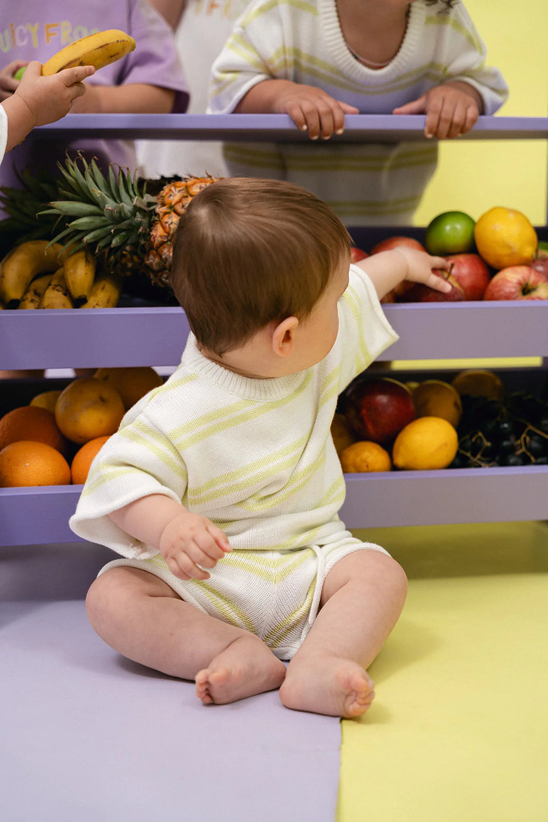 A baby dressed in the Contrast Knit Set Lime/Off White by MYLA JANE sits on the floor, reaching towards a shelf filled with mix-and-match fruits, including apples and oranges. Another person holds a banana near the shelf, all set against a pastel yellow and purple background.