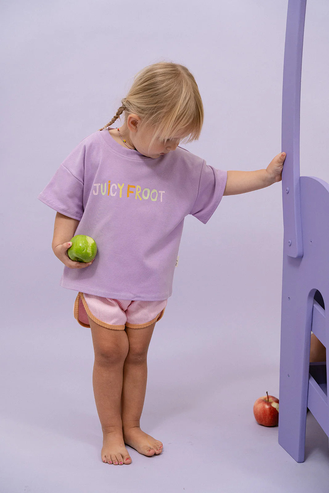 A toddler stands barefoot on a light purple floor, wearing an oversized fit lavender T-shirt with "JUICY FROOT" printed on it. They are holding a green apple in one hand and touching a purple piece of furniture with the other, while MYLA JANE's Contrast Rib Shorts Strawberry complete the playful look. An apple lies nearby.