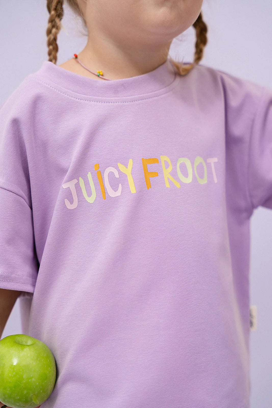 A child wearing an oversized fit lavender Juicy Froot T-Shirt Grape by MYLA JANE, showcasing the vibrant "JUICY FROOT" text, is holding a green apple. This playful yet comfortable tee, made from durable cotton, adds a charming touch to any day.