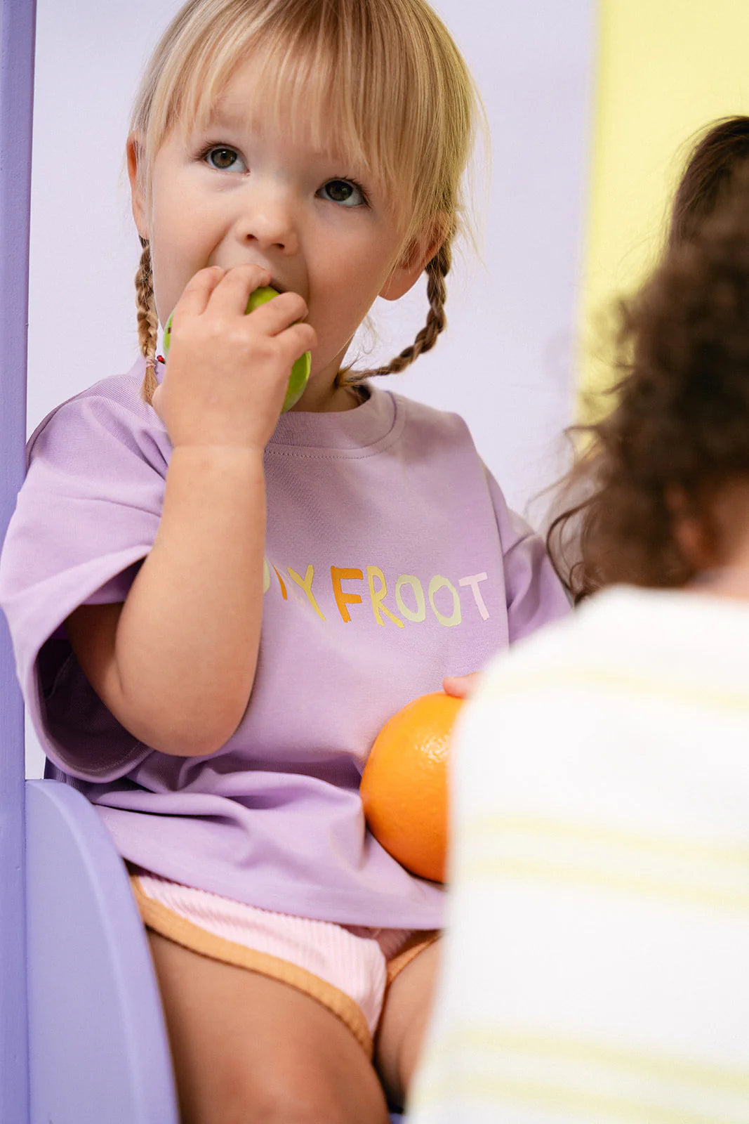 A young child with blonde hair and pigtails is wearing MYLA JANE's Contrast Rib Shorts Strawberry and an oversized purple shirt as they savor a green apple, also holding an orange. The quality cotton rib fabric of the product ensures comfort, while another child's shoulder appears partially in the foreground, enhancing the cozy scene.