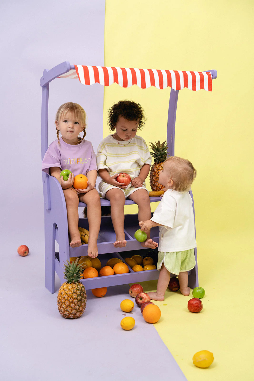 Three toddlers enjoy a lively, market-themed setup. Two are seated on a purple stand, dressed in MYLA JANE's Juicy Froot T-Shirt Grape, holding apples as the third reaches for fruit. The scene is filled with apples, oranges, and pineapples against a cheerful yellow and purple backdrop.