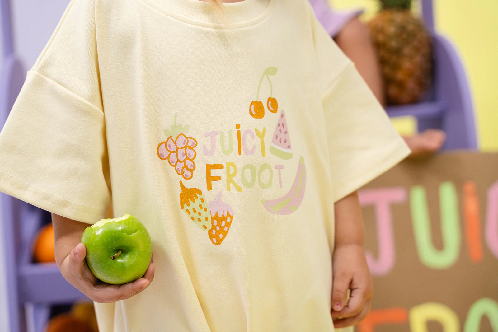 A child wearing an oversized MYLA JANE Juicy Froot T-Shirt Romper Lemon adorned with vibrant fruit illustrations holds a green apple. The durable cotton fabric complements the playful background of colorful fruit-themed decor.