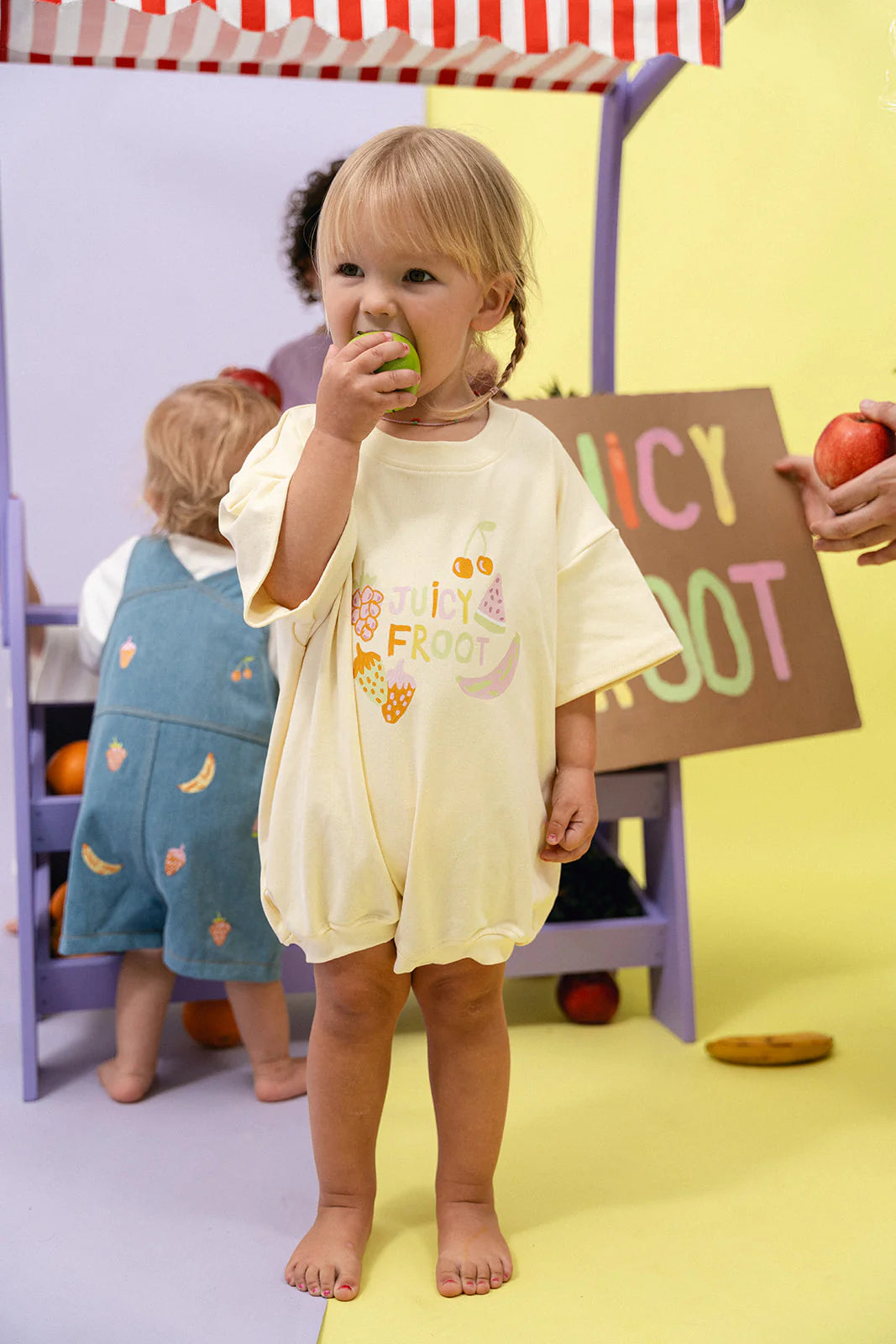 A toddler wearing a Lemon T-Shirt Romper from MYLA JANE's Juicy Froot collection enjoys a green apple. In the background, another child dressed in an oversized fit blue dress with fruit patterns sits under a durable cotton canopy featuring red and white stripes and a "Juicy Froot" sign against a cheerful yellow backdrop.