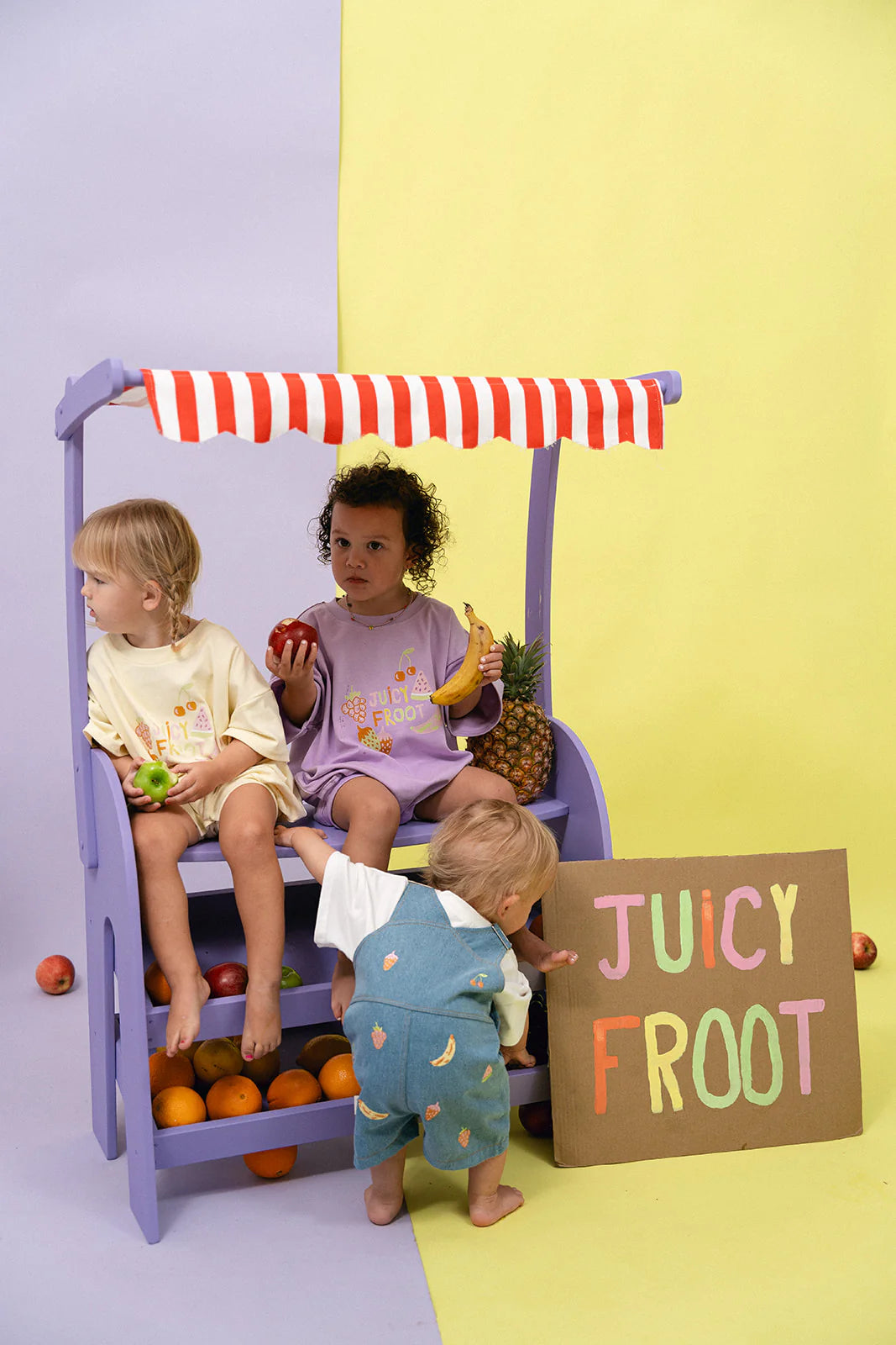 Three children are playing at a vibrant fruit stand. One is holding an apple, and another has a banana. A toddler, wearing the MYLA JANE Juicy Froot T-Shirt Romper Lemon with its oversized fit and durable cotton fabric, reaches for oranges on the stand while a "Juicy Froot" sign is displayed nearby. The background features a blend of pastel purple and yellow.