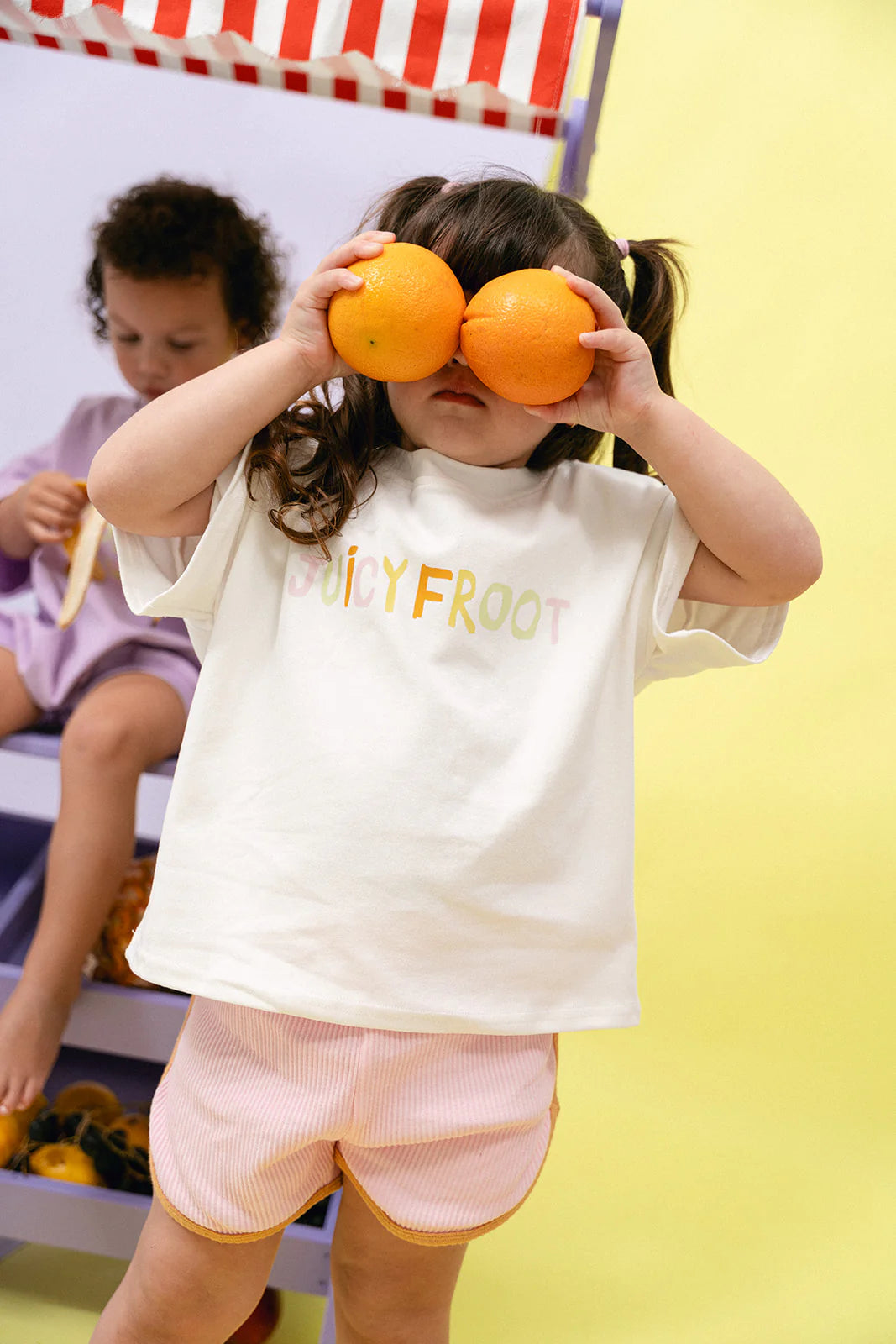 A young girl in an oversized fit MYLA JANE's "Contrast Rib Shorts Strawberry" playfully holds two oranges over her eyes. She stands in front of a fruit stand with another child sitting in the background, both surrounded by bright colors and fruits.