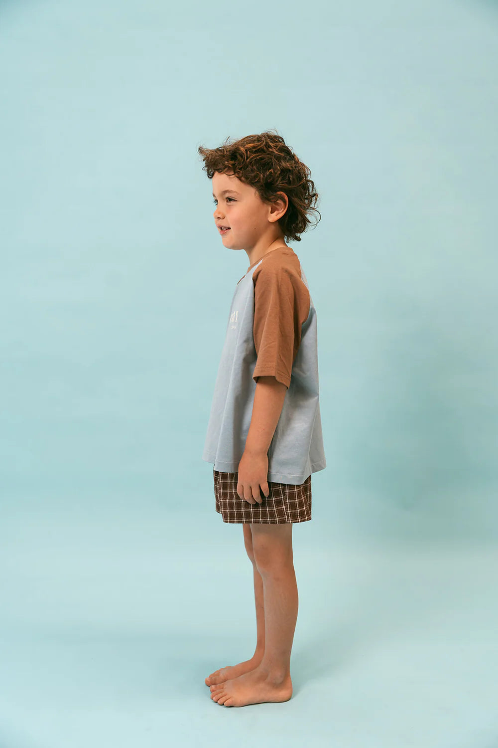 A young child stands barefoot in profile against a light blue background, wearing the Howdy Raglan Tee by LENN LABEL with relaxed fit brown checkered shorts. The Cotton Jersey fabric complements the child's curly hair as they face left.