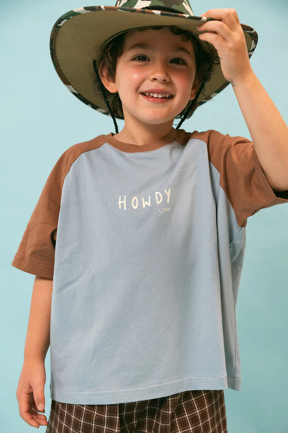 A smiling child wears a large camo hat and the Howdy Raglan Tee by LENN LABEL, featuring "HOWDY" in offset print. The tee has blue and brown sleeves that perfectly match the brown plaid pants, all made from Cotton Jersey. This vibrant ensemble stands out against a solid light blue background.