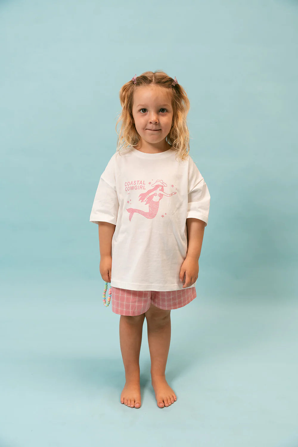 A young child with wavy blonde hair stands smiling in front of a light blue background. They are wearing the LENN LABEL Coastal Cowgirl Tee, a relaxed fit white Cotton Jersey T-shirt featuring a pink mermaid graphic and the phrase "Coastal Cowgirl," paired with pink checkered shorts for an easy-going style.