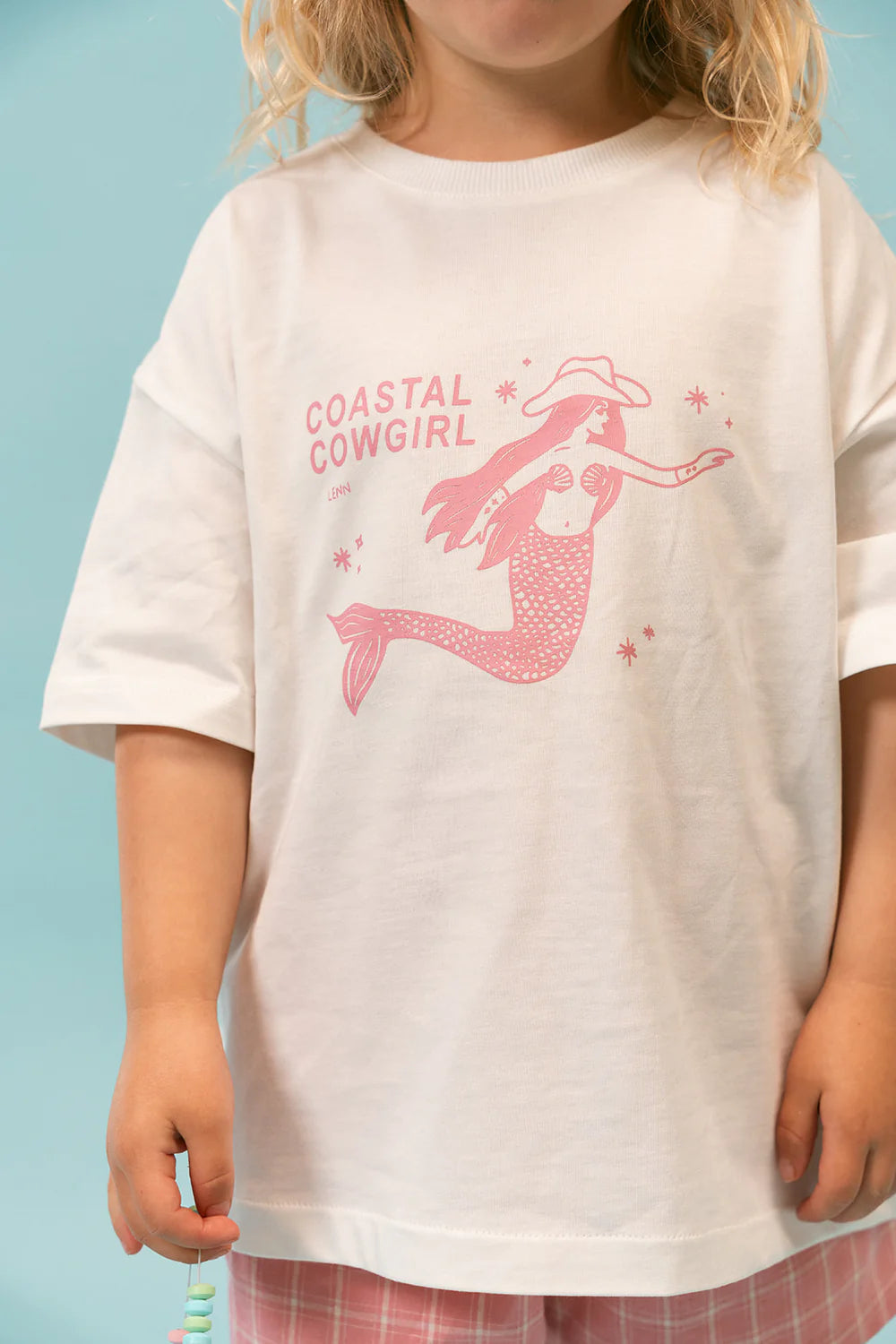 A child showcases the Coastal Cowgirl Tee by LENN LABEL, featuring a whimsical design of a pink mermaid wearing a cowboy hat and boots on a white cotton jersey. The relaxed fit is accentuated by the light blue background, perfectly paired with plaid shorts for a fun and playful look.