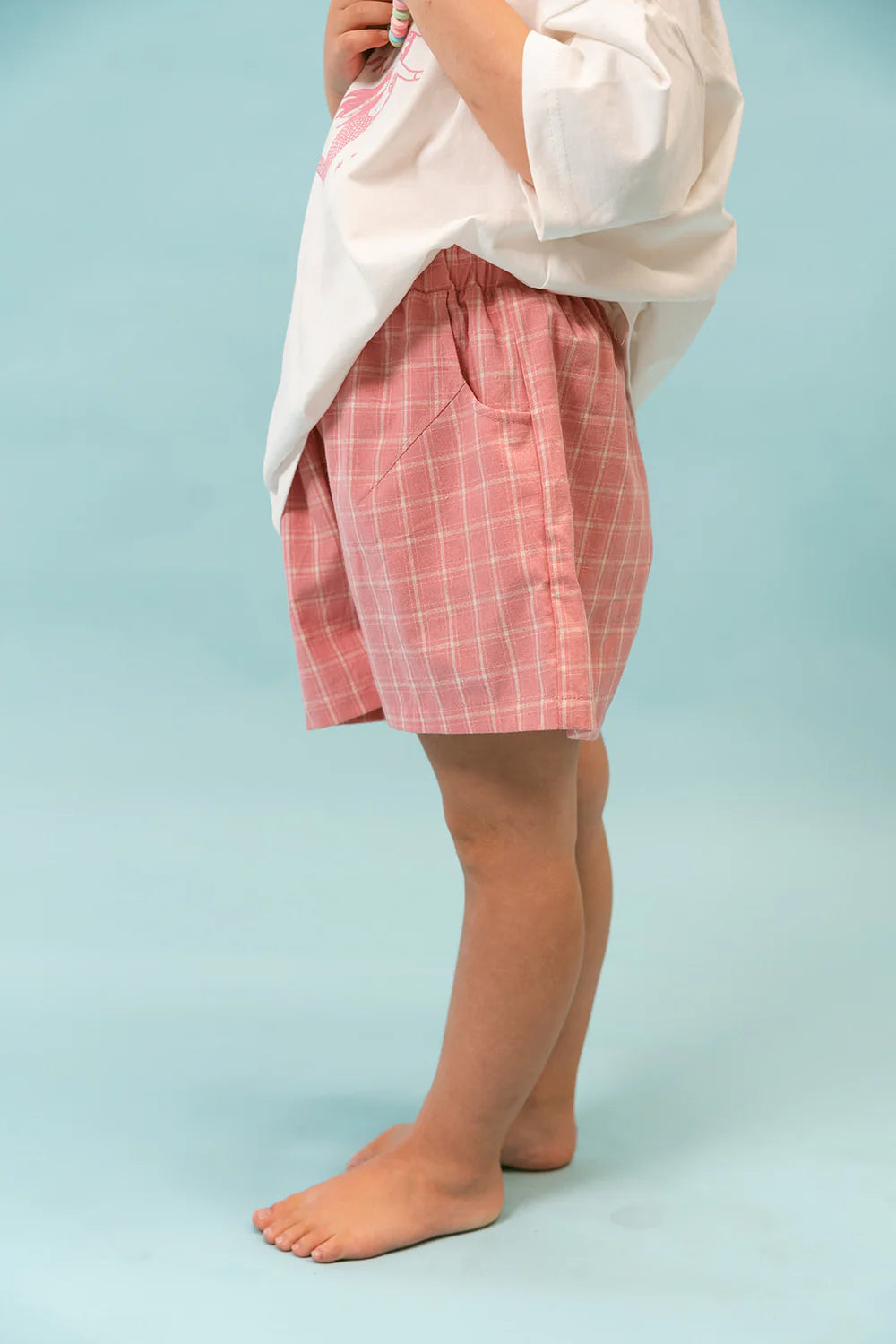 A child stands barefoot against a light blue background, wearing a white cotton shirt and Check Short Lemonade with an elastic waistband by LENN LABEL.