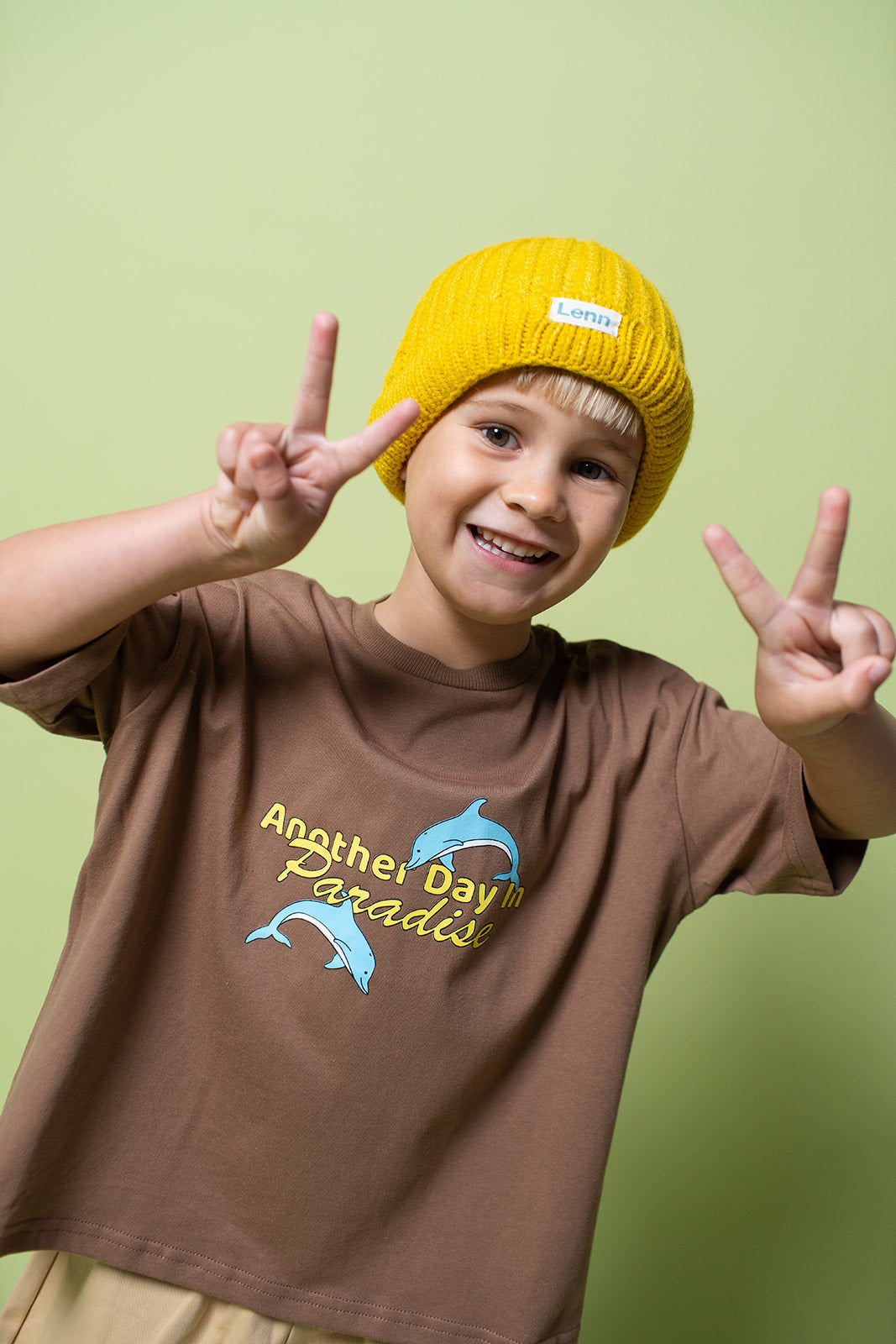 A young kid wearing the Lenn paradise tee in brown with a yellow beanie on making peace signs with his hands.