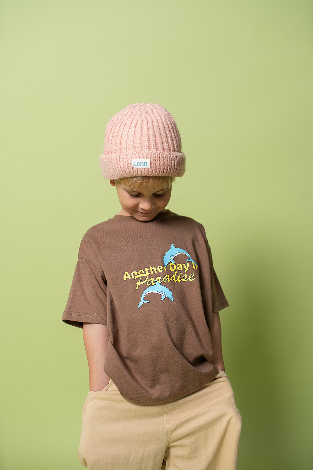 A young kid wearing the Lenn paradise tee  in brown with a peach beanie and mustard pants on.