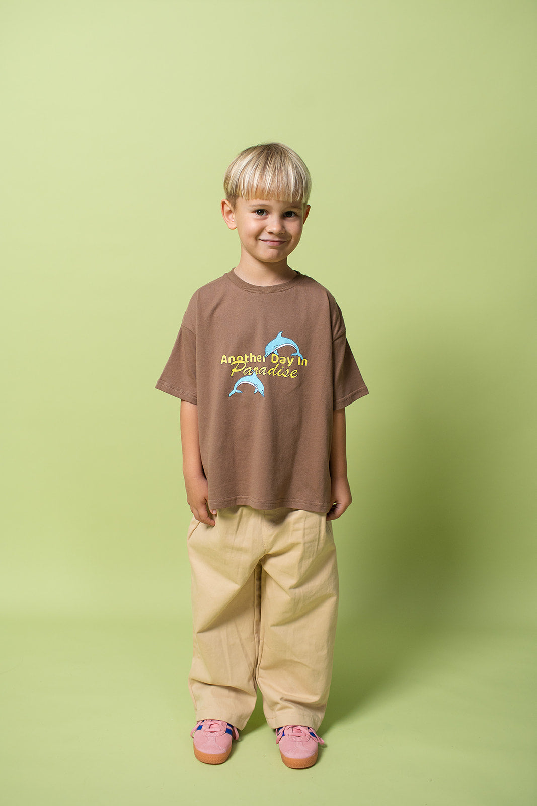 A young kid wearing the Lenn paradise tee  in brown with mustard pants on and pink shoes.