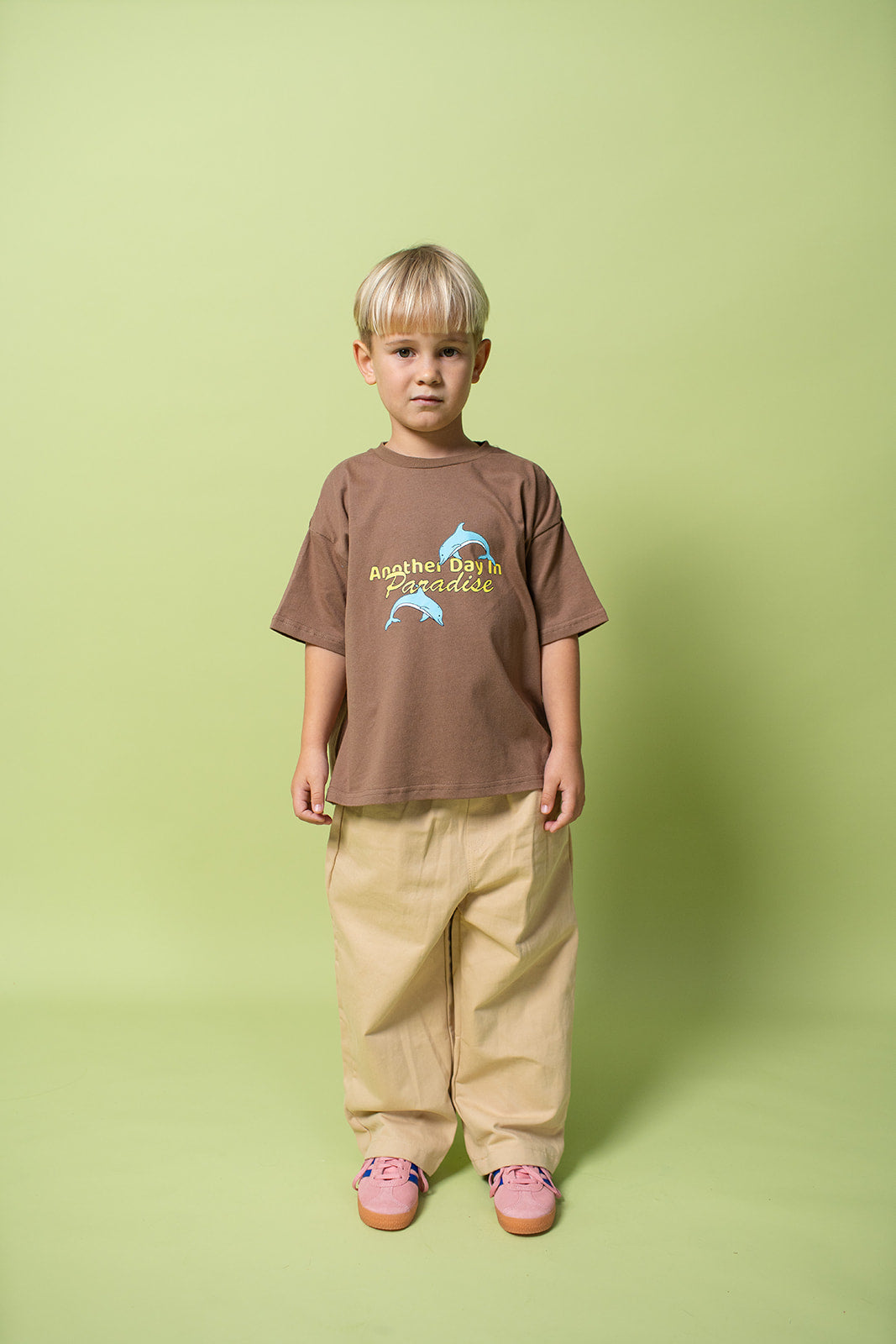 A young kid wearing the Lenn paradise tee  in brown with mustard pants on and pink shoes.