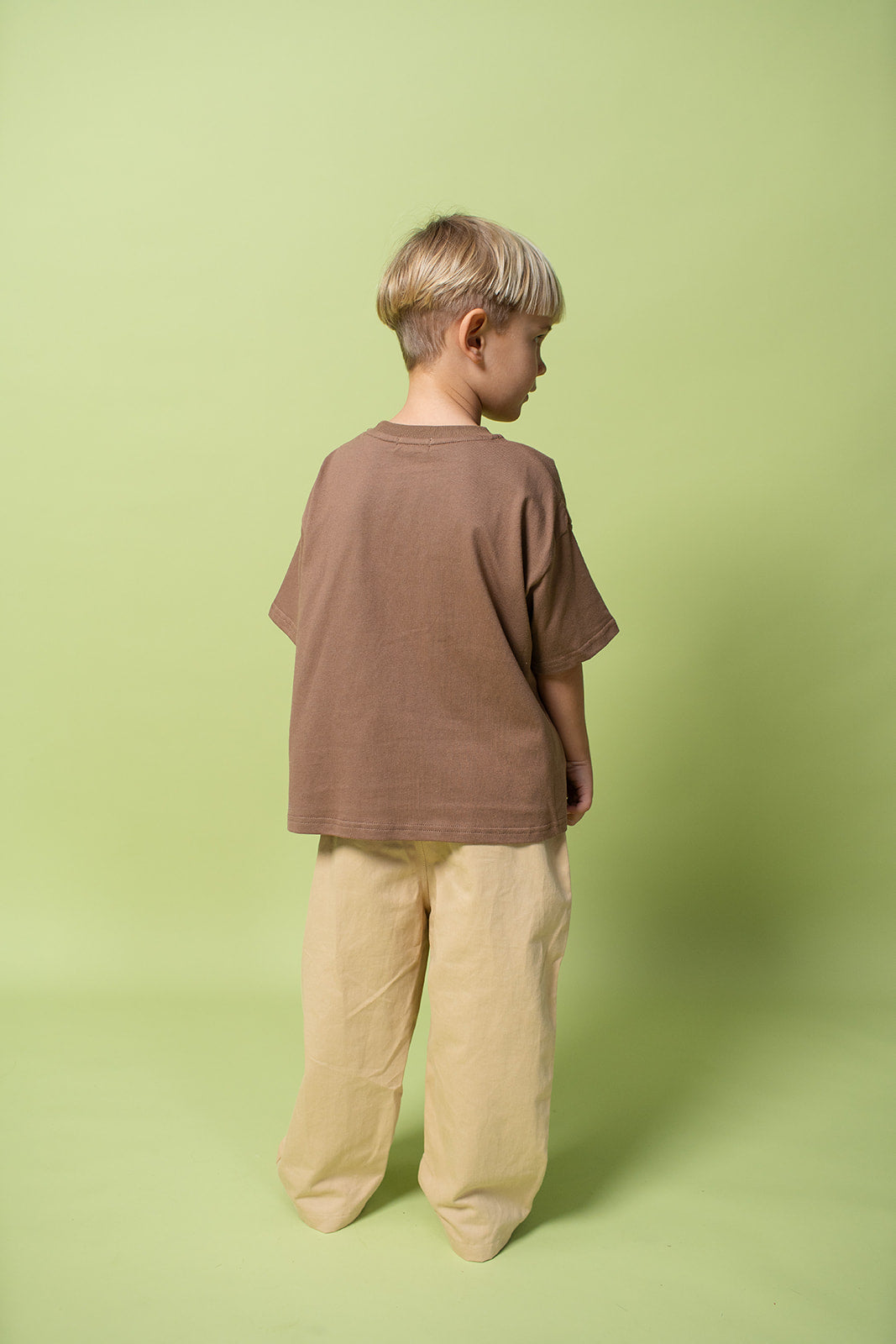 A young kid wearing the Lenn paradise tee  in brown with mustard pants on from the back.