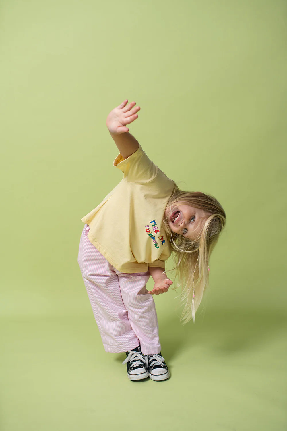 A young child with long blonde hair, wearing an oversized fit "Rainbow Tee" from LENN LABEL, made from 100% cotton jersey, paired with pink and white striped pants and black sneakers, playfully bends to the side and waves with one hand against a light green background.