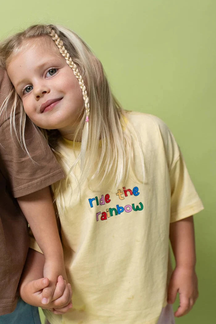 A young girl with long blonde hair, partially braided, is smiling and leaning on the shoulder of someone next to her. She is wearing an oversized, light yellow 100% cotton LENN LABEL Rainbow Tee featuring colorful lettering that reads "ride the rainbow." The background is a light green color.