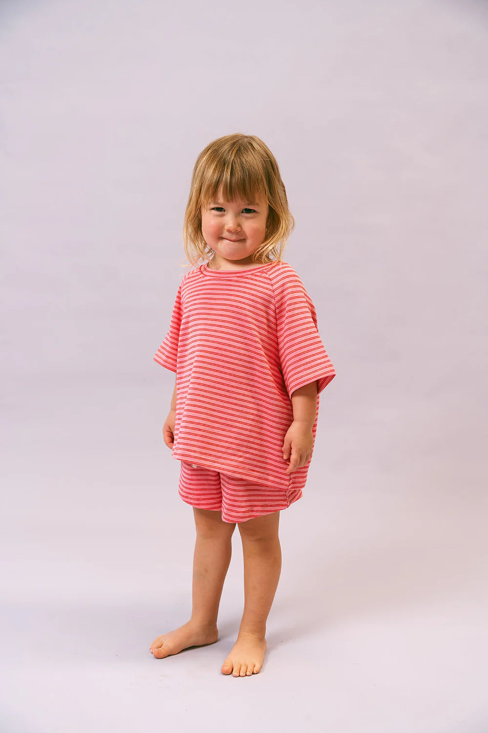 A young child with blonde hair wears the oversized Lenn Label ~ Sunday Set Watermelon by LENN LABEL, featuring a pink and white striped shirt and shorts with an elastic waistband. Standing barefoot against a light gray backdrop, the child smiles slightly, facing forward with hands by their sides.