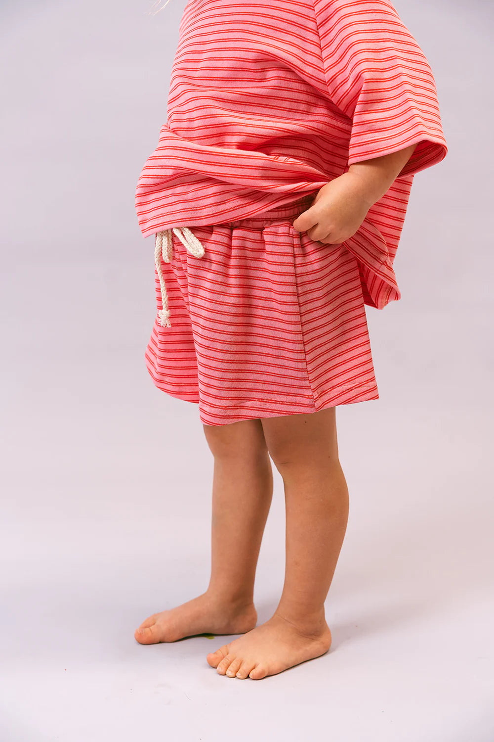 A child models the Lenn Label ~ Sunday Set Watermelon by LENN LABEL, a red and white striped cotton outfit with loose shorts featuring an elastic waistband and a matching oversized top. The barefoot child poses relaxedly against a light background.