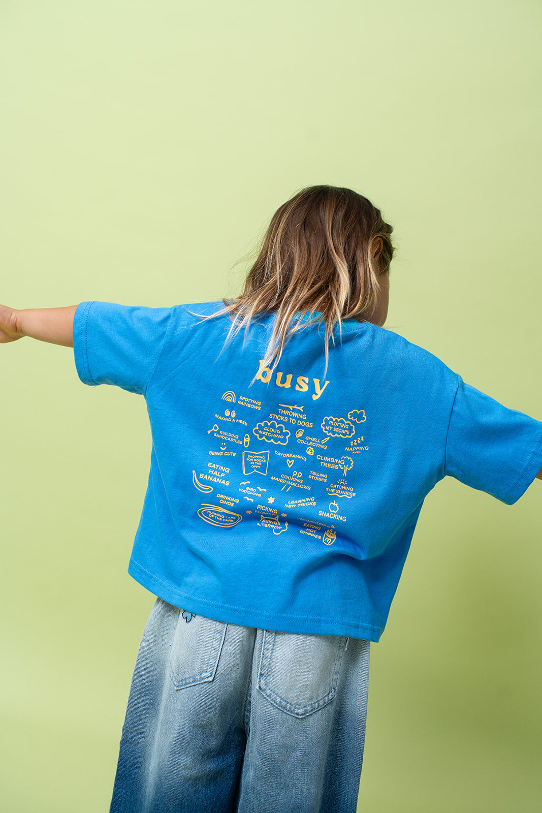 A young kid wearing the Lenn busy tee sunny shirt in blue from the back with their arms out stretched.