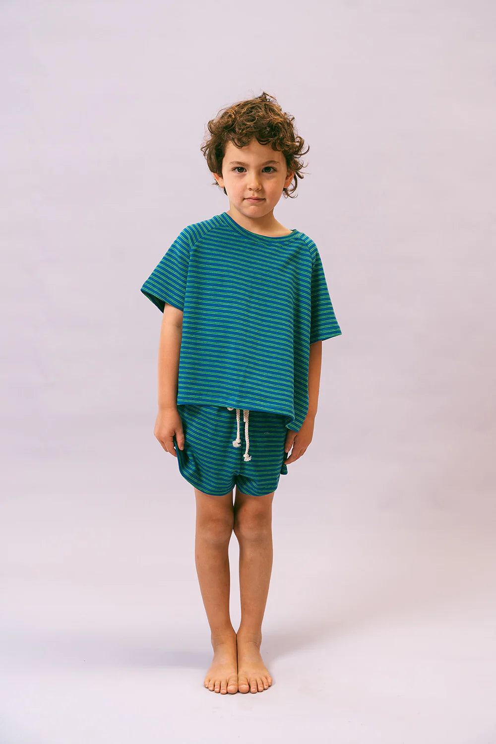 A young child with curly hair stands barefoot against a plain background, wearing the Lenn Label ~ Sunday Set Sea. This oversized, elastic waistband set from LENN LABEL features a blue and green striped stretch cotton T-shirt and matching shorts. The child appears calm, with their hands at their sides.