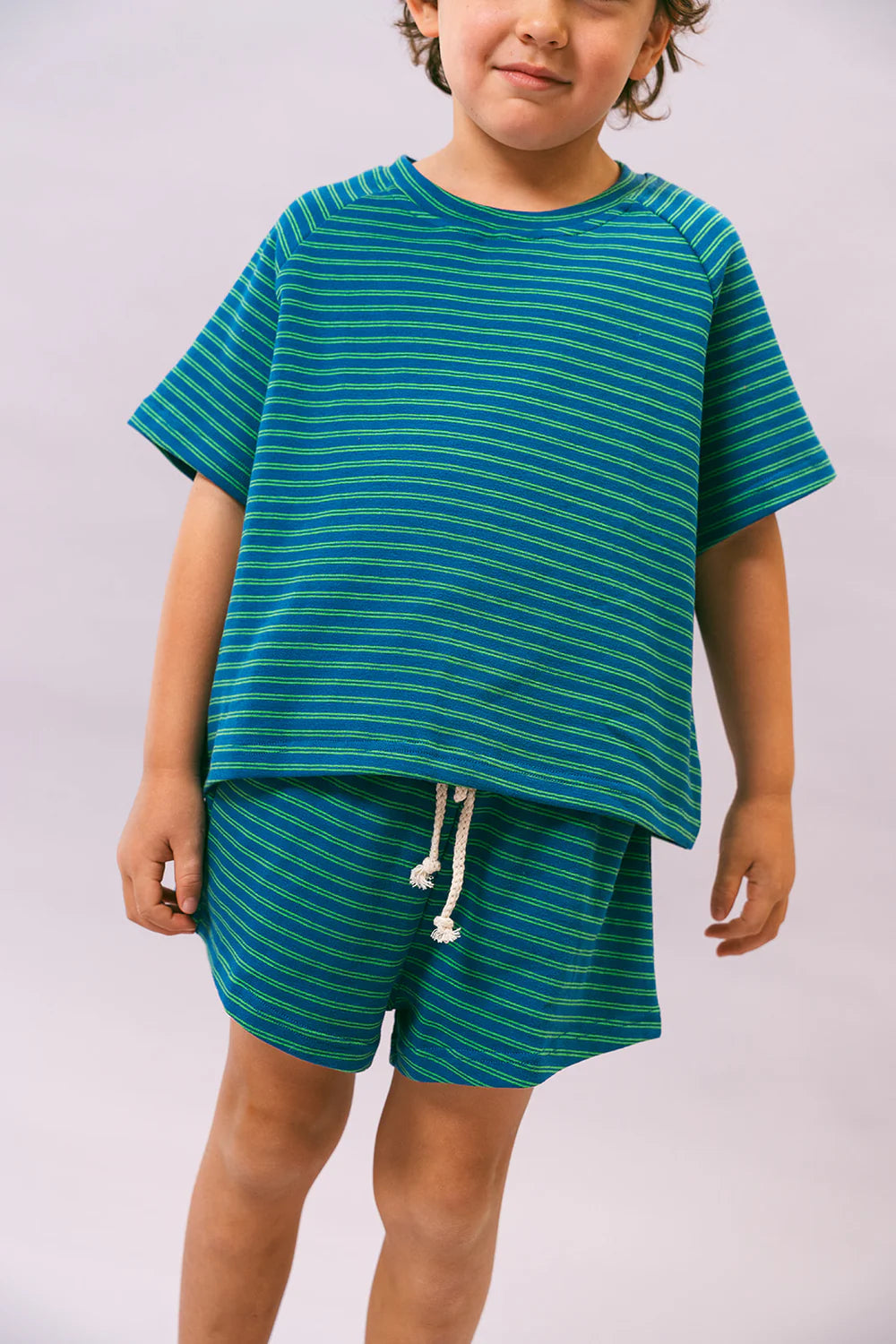 A child wears the Lenn Label ~ Sunday Set Sea from LENN LABEL, featuring an oversized blue and green striped t-shirt and matching shorts with an elastic waistband, made of stretch cotton for comfort against a plain, light background.