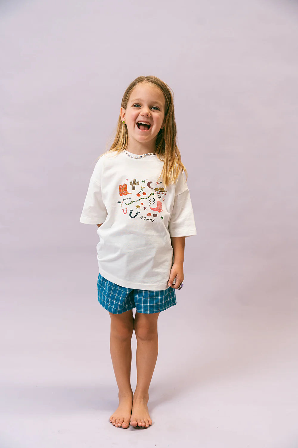 A smiling child with long, light brown hair stands barefoot against a plain background, showcasing a relaxed fit in the LENN LABEL Cowboy Tee, a white cotton jersey T-shirt featuring colorful designs, paired with blue plaid shorts.