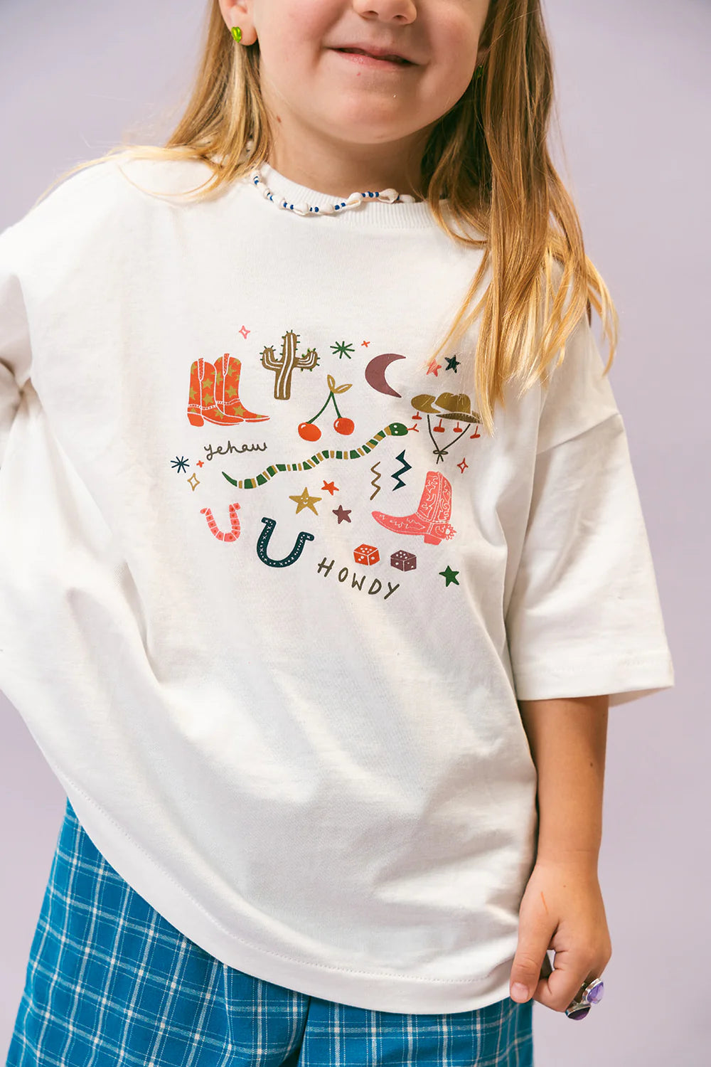 A child is wearing the Cowboy Tee by LENN LABEL, an oversized cotton jersey T-shirt adorned with vibrant Western-themed illustrations such as boots, cacti, and a snake, alongside the words "Yeehaw" and "Howdy." The outfit is complemented by blue plaid pants and a beaded necklace, creating a playful and relaxed look.