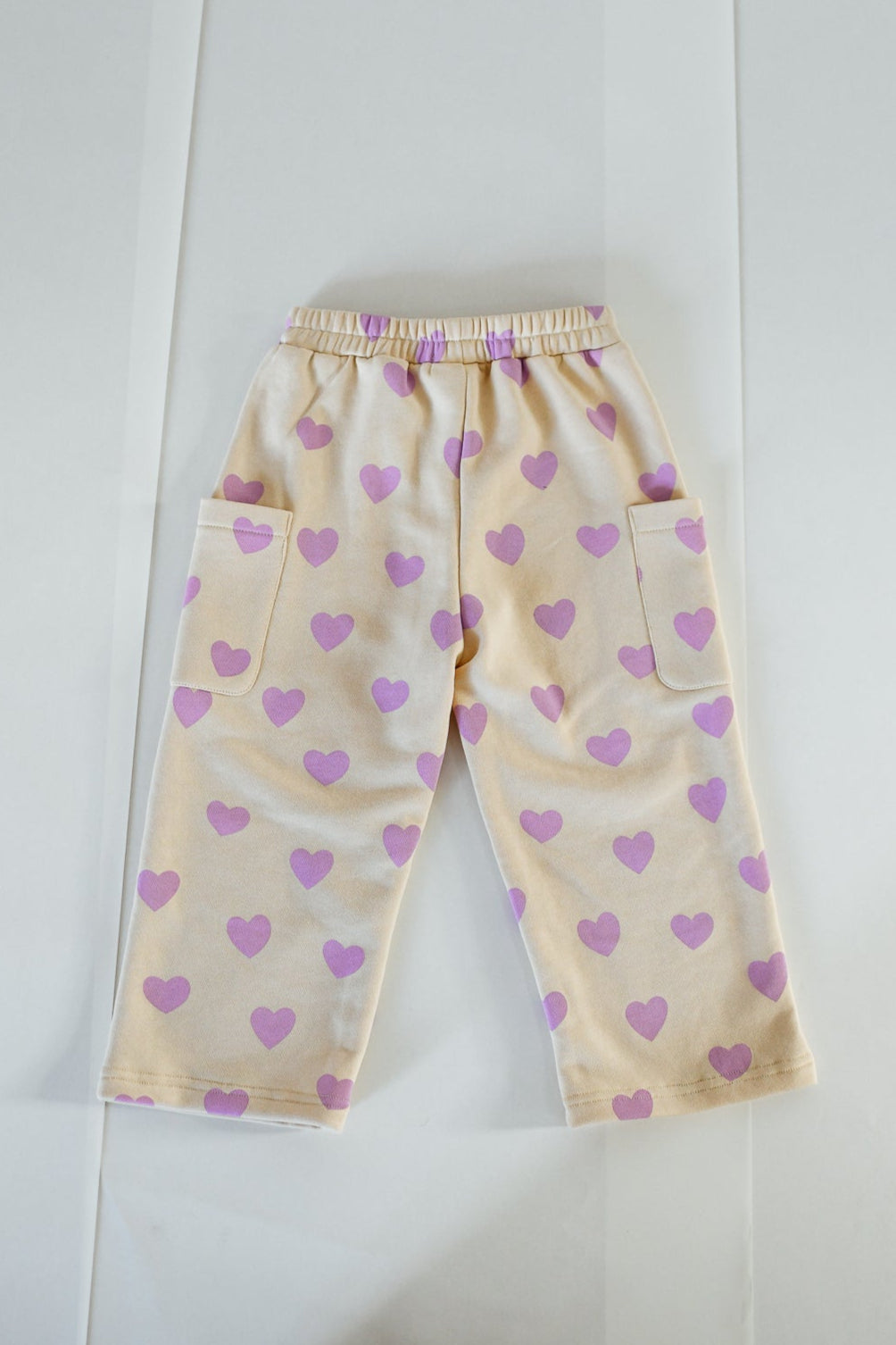 The SUNDAY SIBLINGS Sunday Siblings - Holiday Pants Purple Hearts are kids' beige french terry cotton pants with an elastic waistband, wide legs, large side pockets, and a fun purple heart pattern. Laid flat on a white surface.