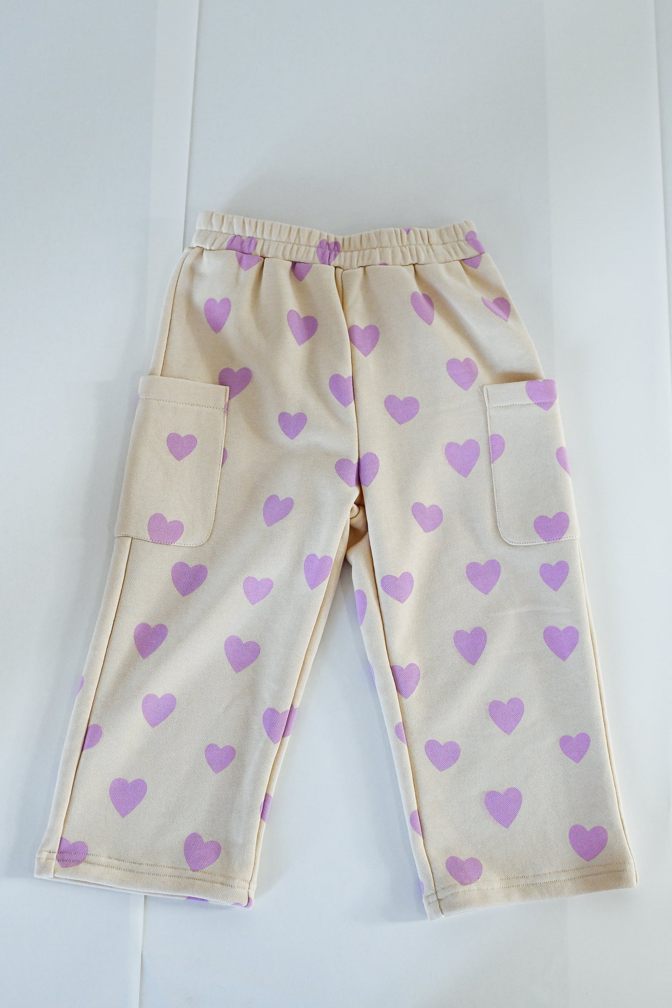 The SUNDAY SIBLINGS Sunday Siblings - Holiday Pants Purple Hearts are beige french terry cotton pants with an elastic waistband, large side pockets, and a playful purple heart pattern, shown laid flat on a white surface.