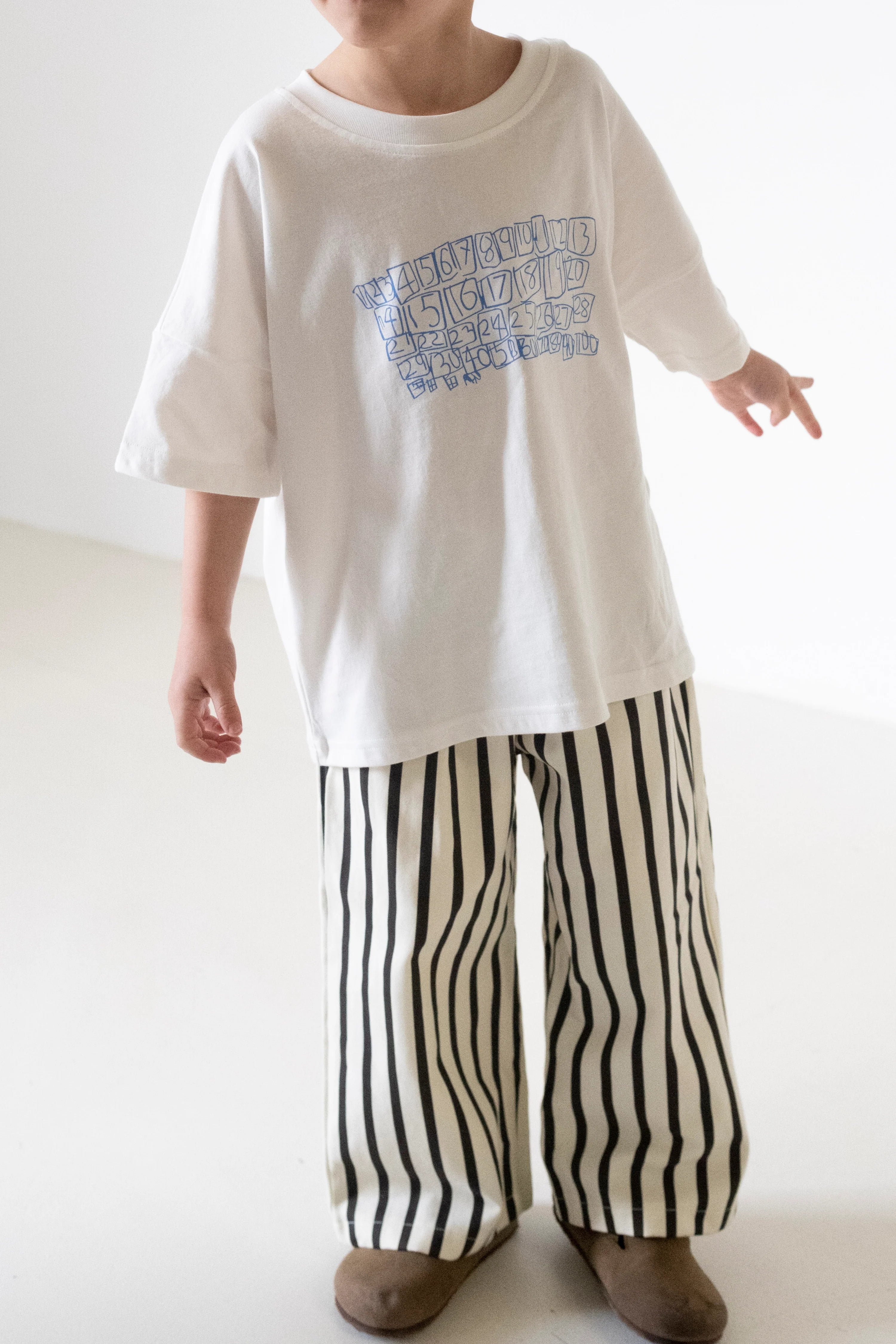 A child wears the TINY TROVE Parker Numbers Relaxed Tee in white/blue, paired with wide-legged white pants with black stripes and brown shoes, standing against a plain white background.