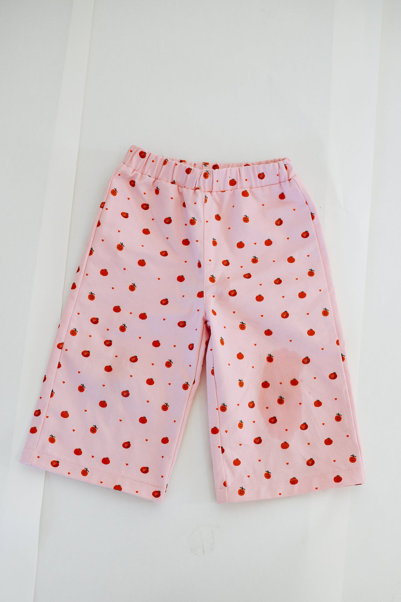 PRE-ORDER SUNDAY SIBLINGS Fruit Loop Pants Pink for kids, made from canvas cotton with an elastic waistband and wide legs. Features a playful red apple print. Shown laid flat on a white background.