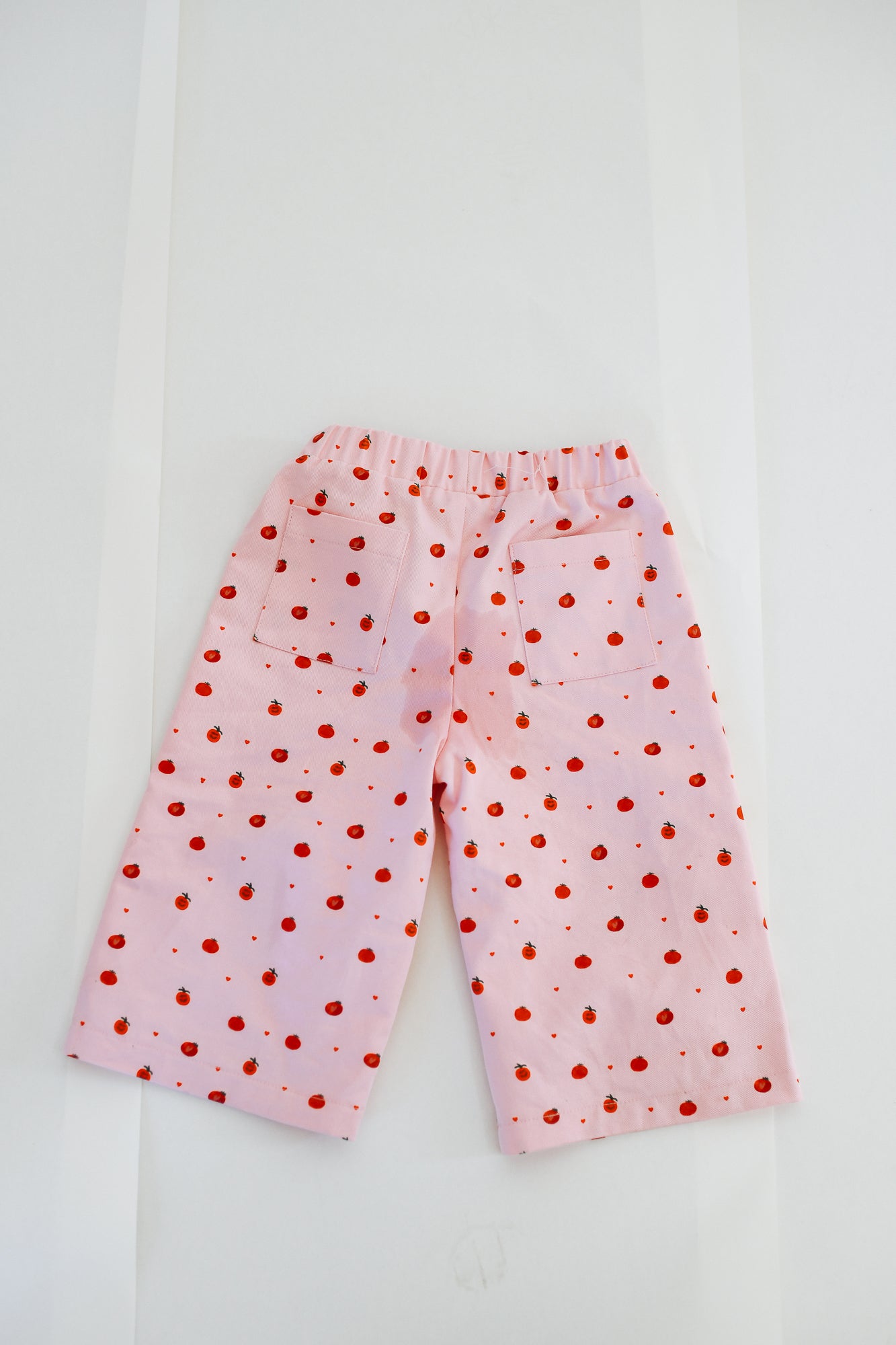 The Sunday Siblings - Fruit Loop Pants Pink by SUNDAY SIBLINGS are light pink canvas cotton pants with red polka dots and two back pockets, shown laid flat on a white background.