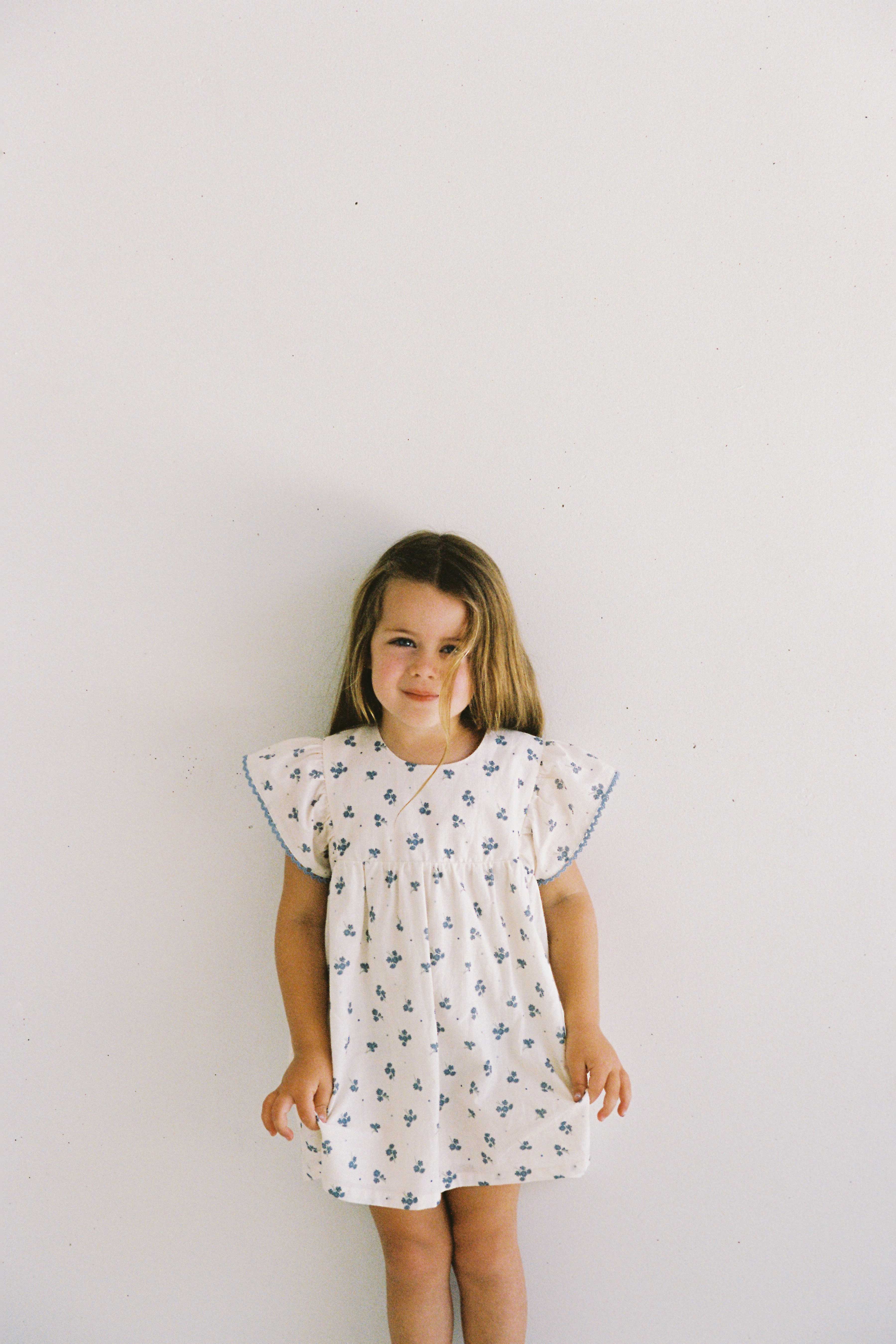 A young girl with long blonde hair stands against a plain white background, wearing the Daisy Dress Ditsy Floral by JUNI JUNIOR. Her dress, made of textured cotton fabric, features blue floral patterns and ruffled sleeves with lace trim detail. She gazes slightly to her left, sporting a subtle smile.